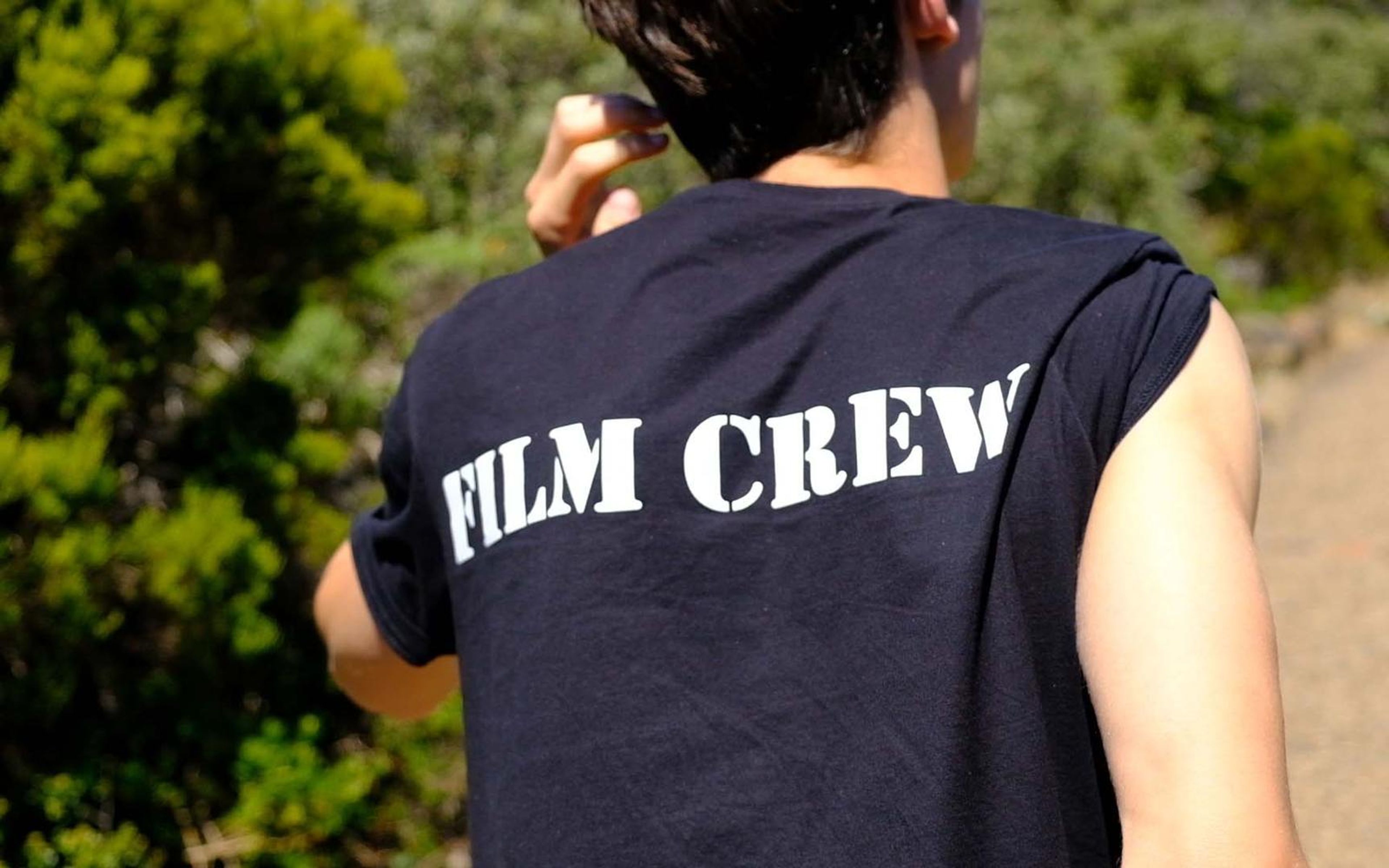 Child in a black sleeveless shirt printed FILM CREW seen from behind against green bush