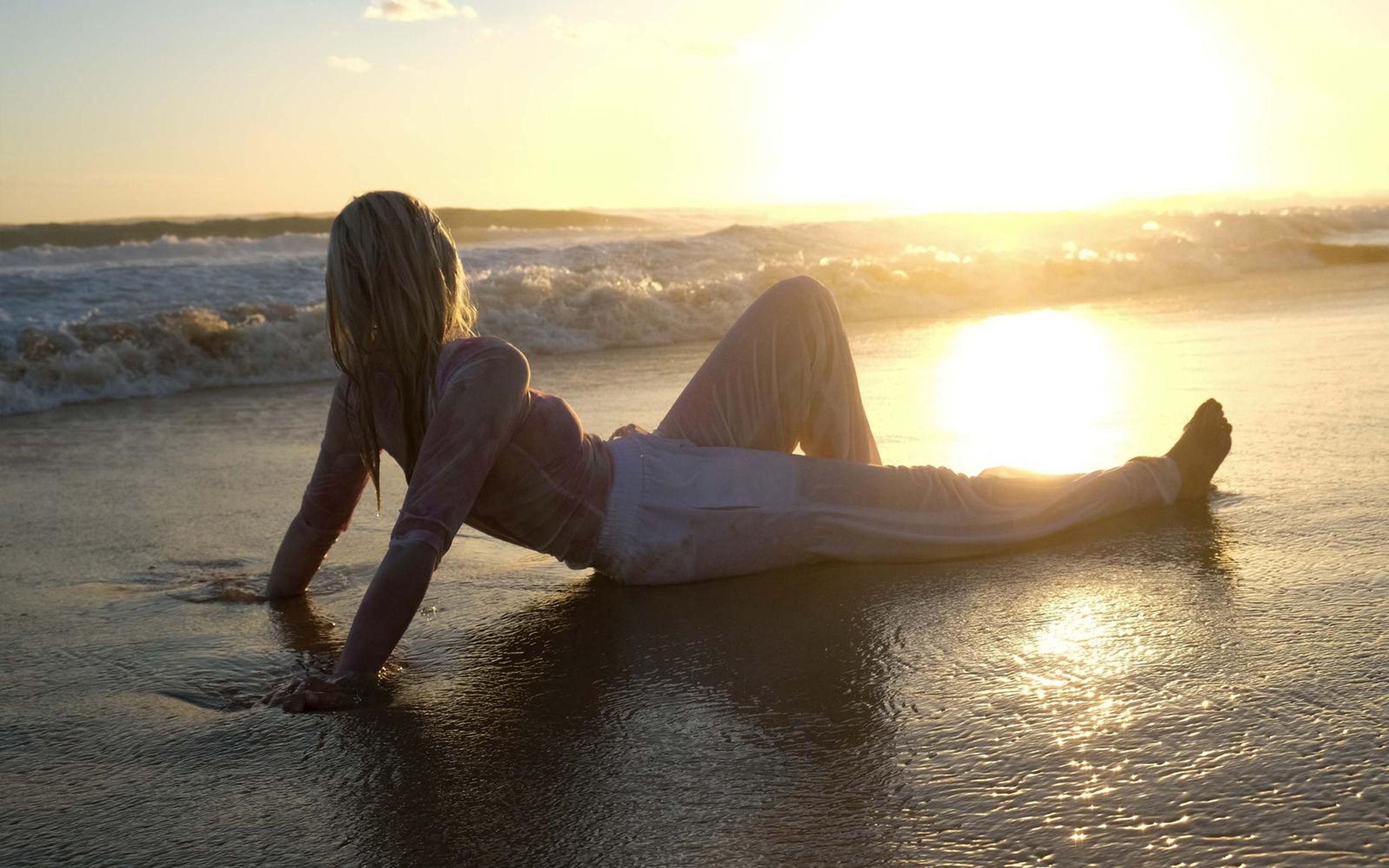 Woman lying on wet sand in a pink patterned top and white trousers, propped on her arms, sun low over the surf