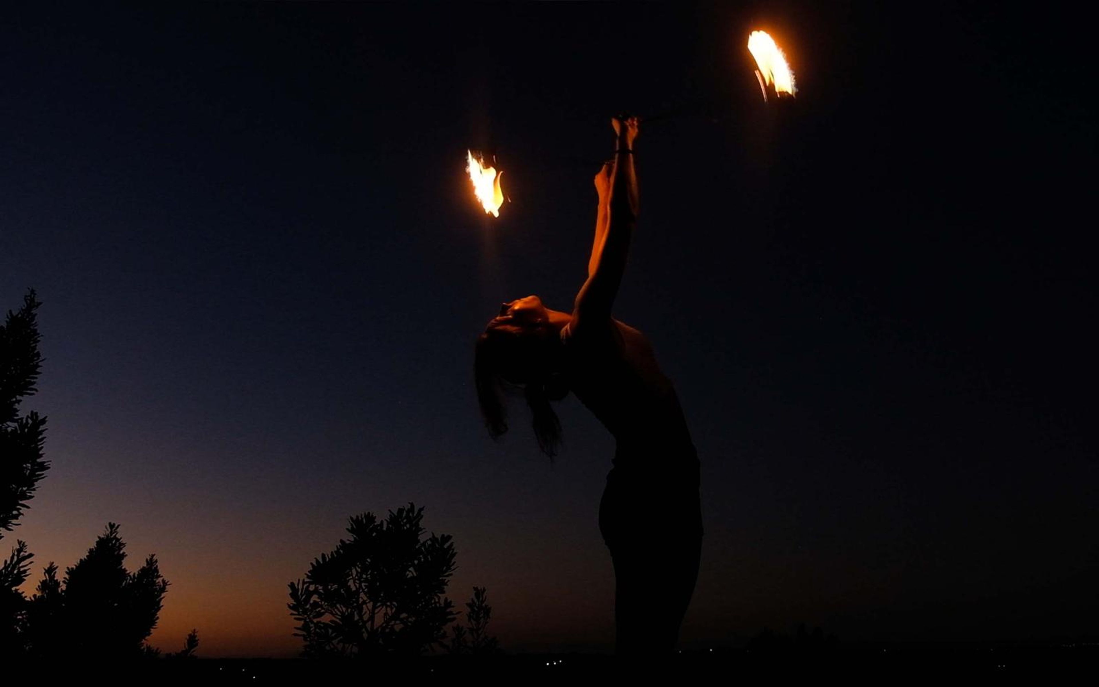 Silhouette of a performer arching back with a fire prop held high against a dusk sky and trees