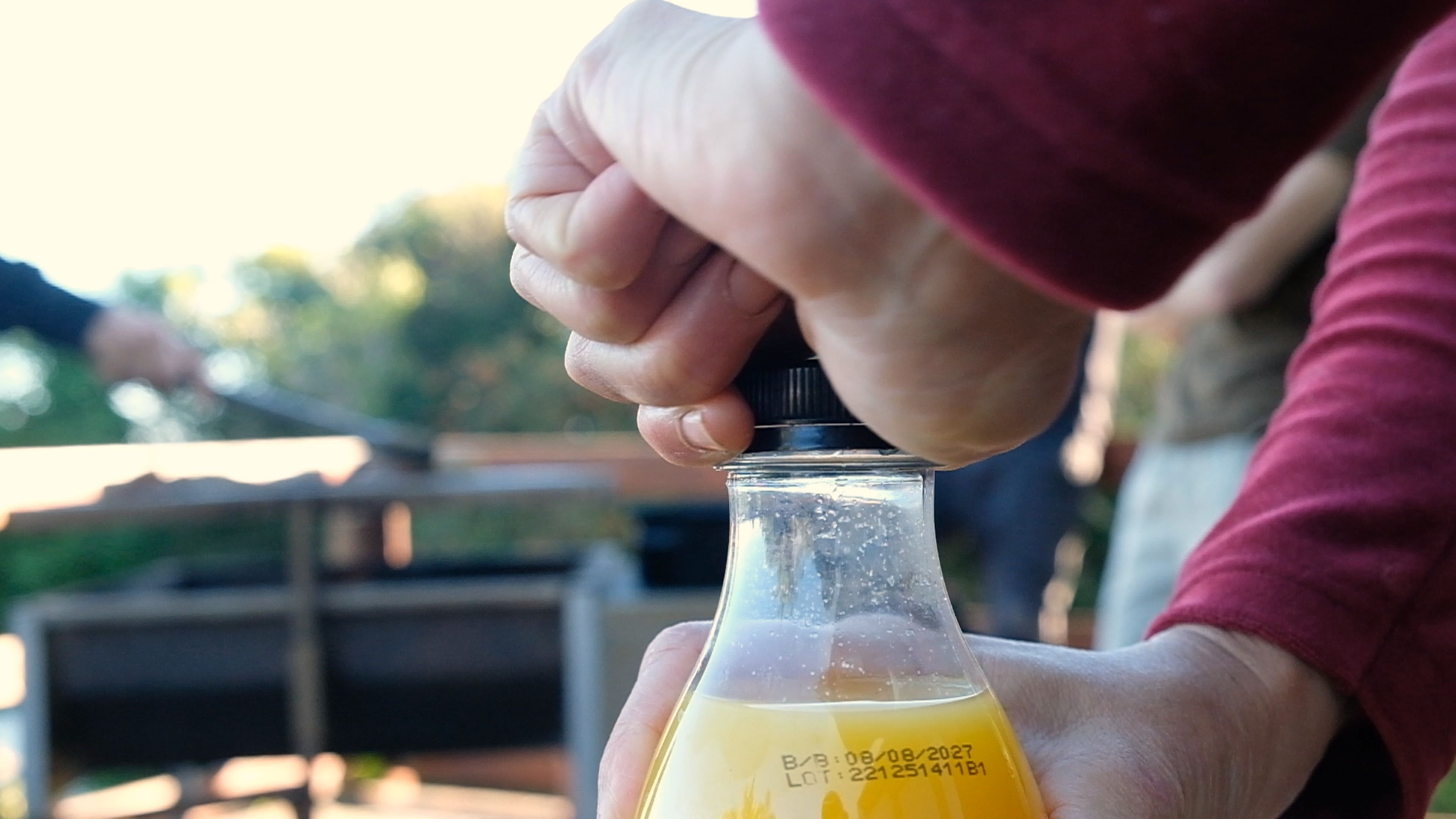Hand twisting the cap off a bottle of orange juice at an outdoor braai
