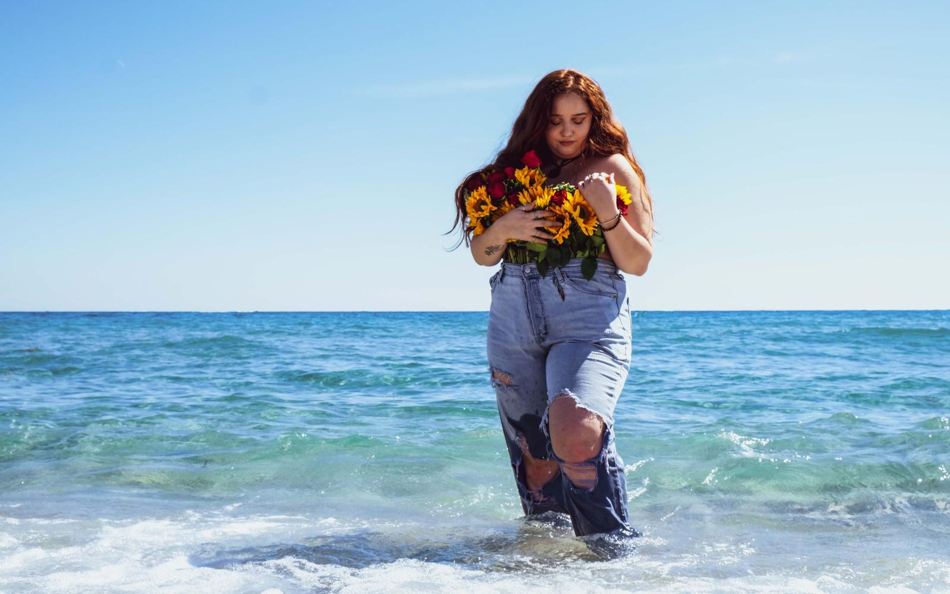 Model wading in a softer pale blue colour grade, bouquet at chest, horizon behind