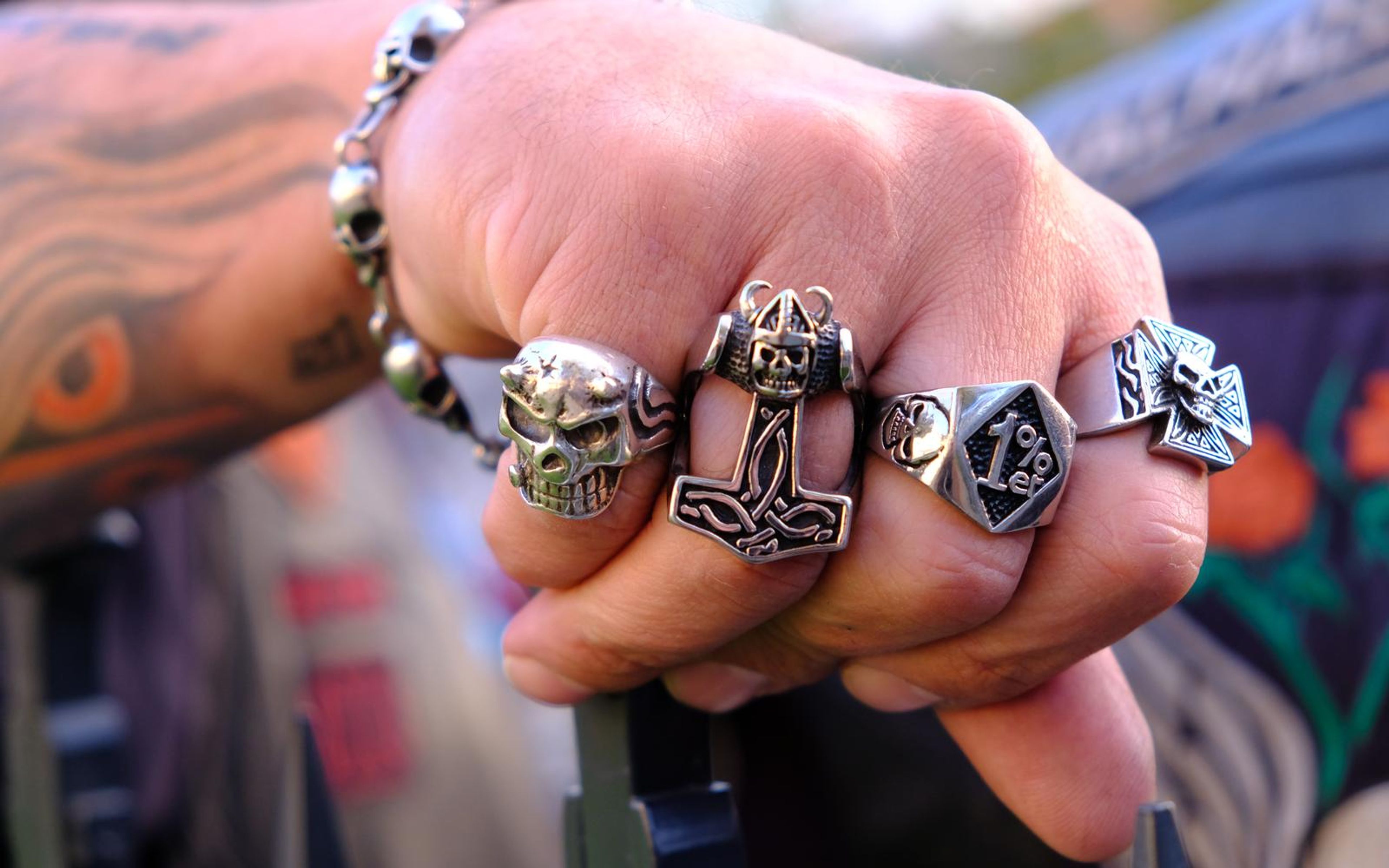 Tattooed hand with four silver skull and cross rings resting on a handlebar, blurred leather vests behind