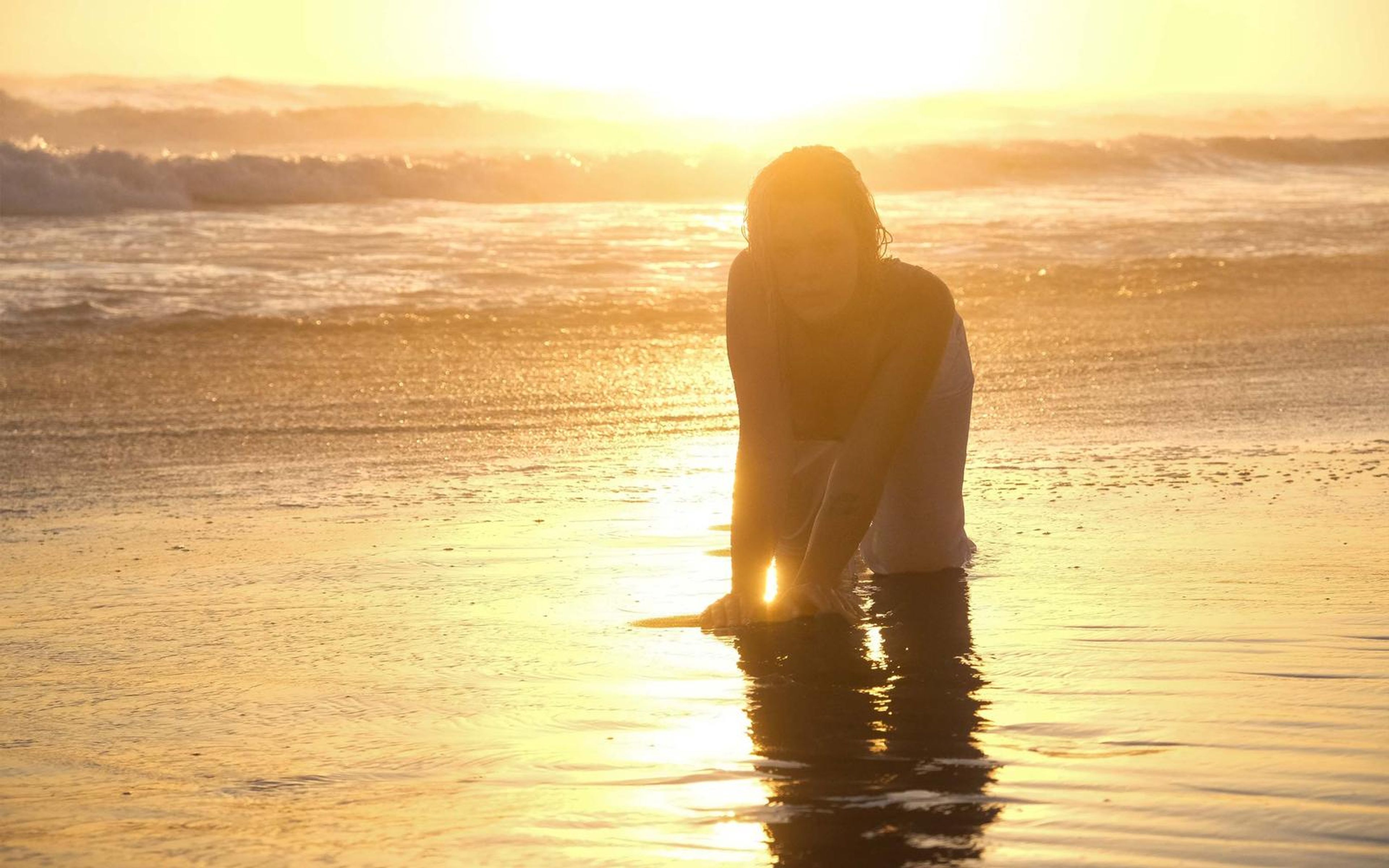 Woman rising from knee-deep water in strong backlight, silhouetted against a sun-streaked shoreline