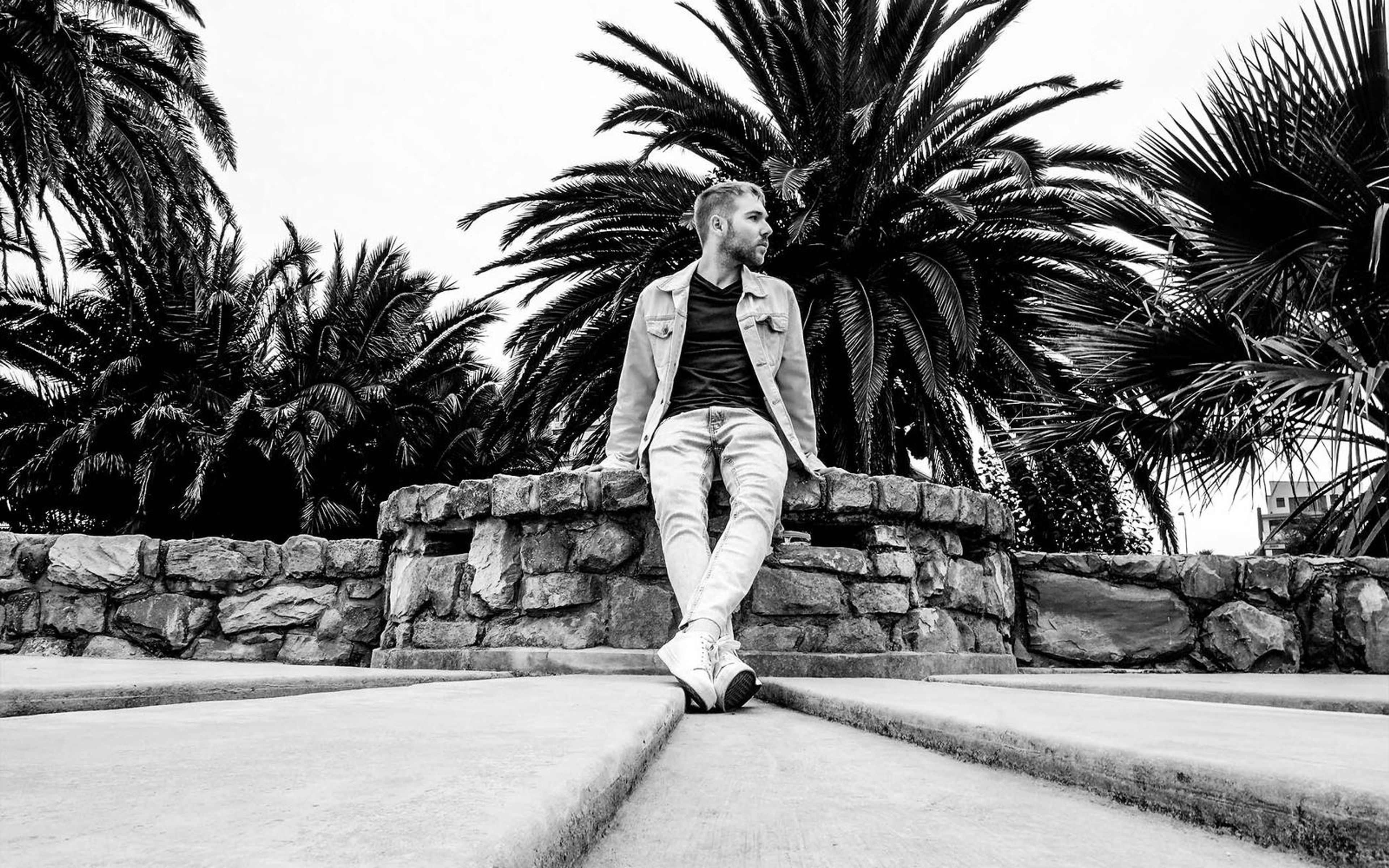 Black and white version of a man sitting on a stone wall, palm trees against a bright sky