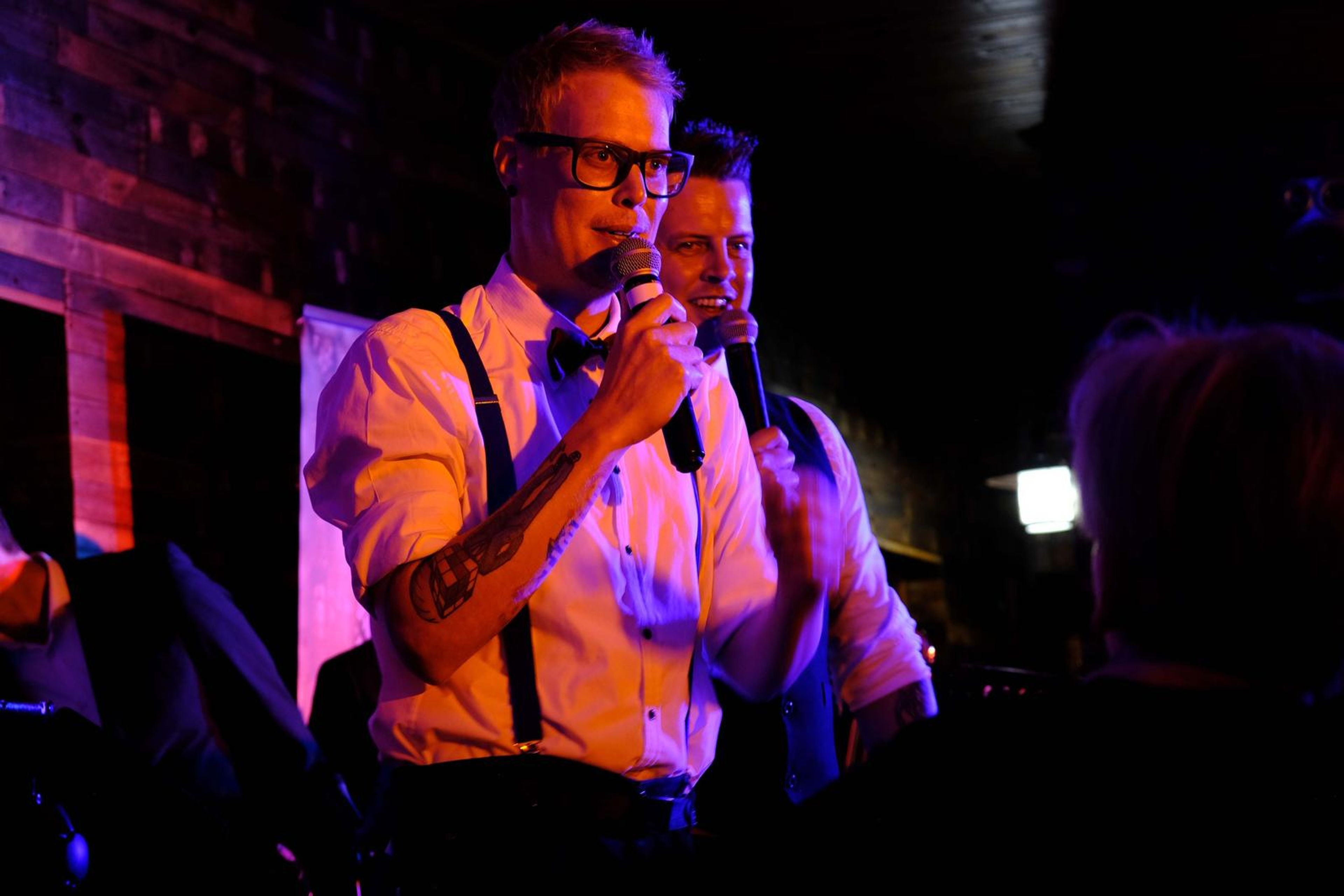 Singer in suspenders and a bow tie singing into the microphone with a bandmate