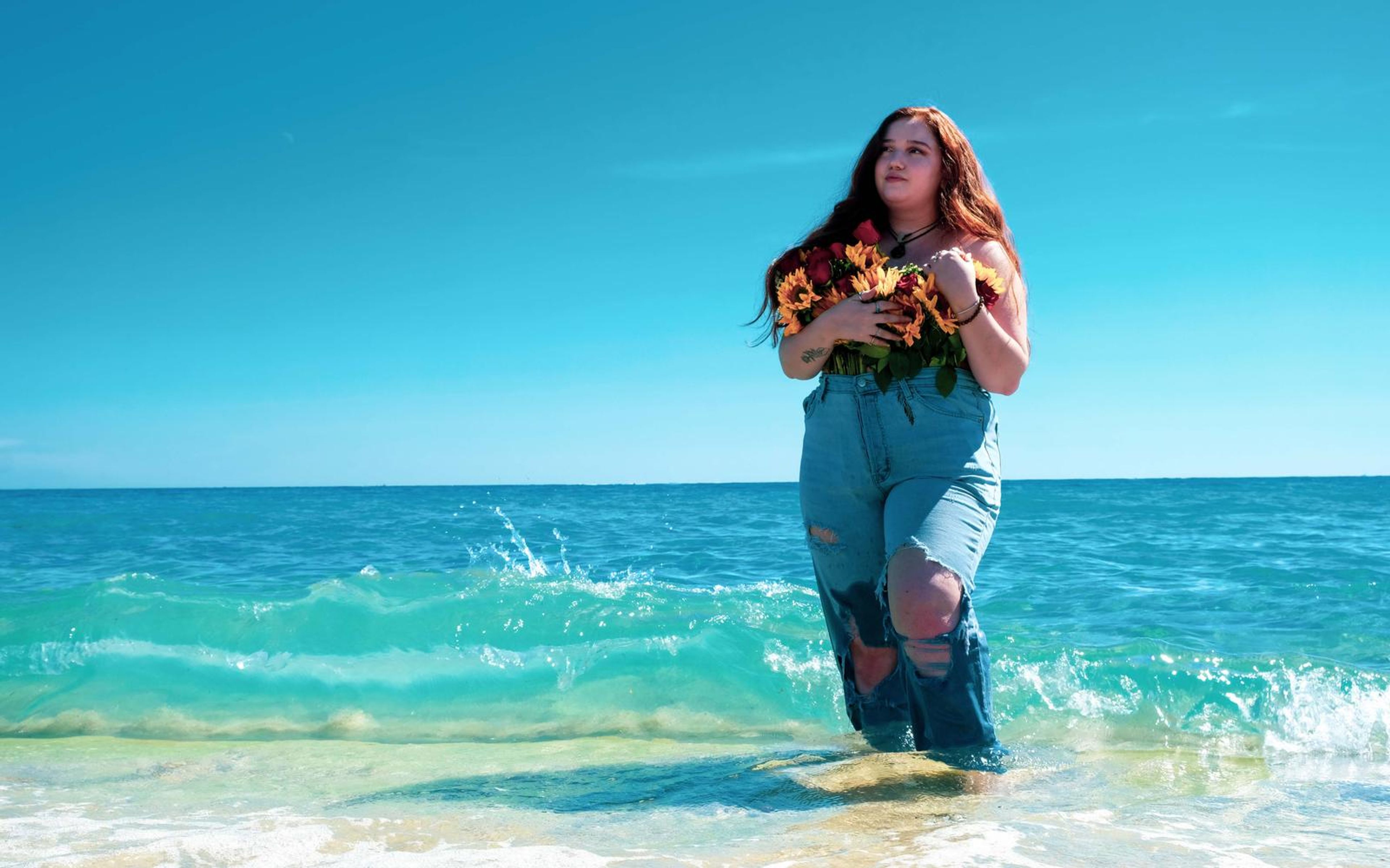 Model standing in clear green-blue shallows holding the bouquet, wet sand in the foreground