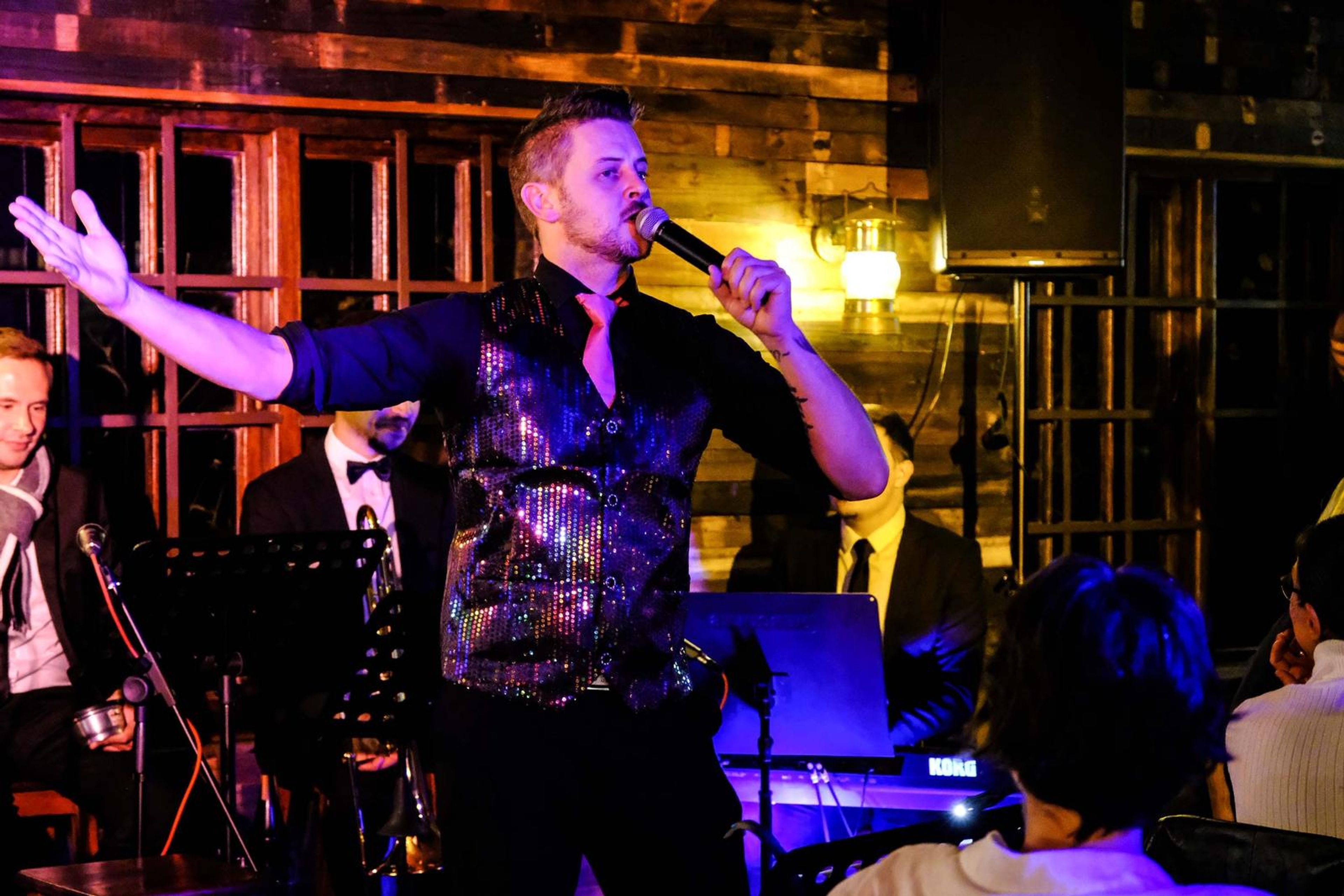 Lead singer in a sequin vest with his arm stretched out as the band plays behind him
