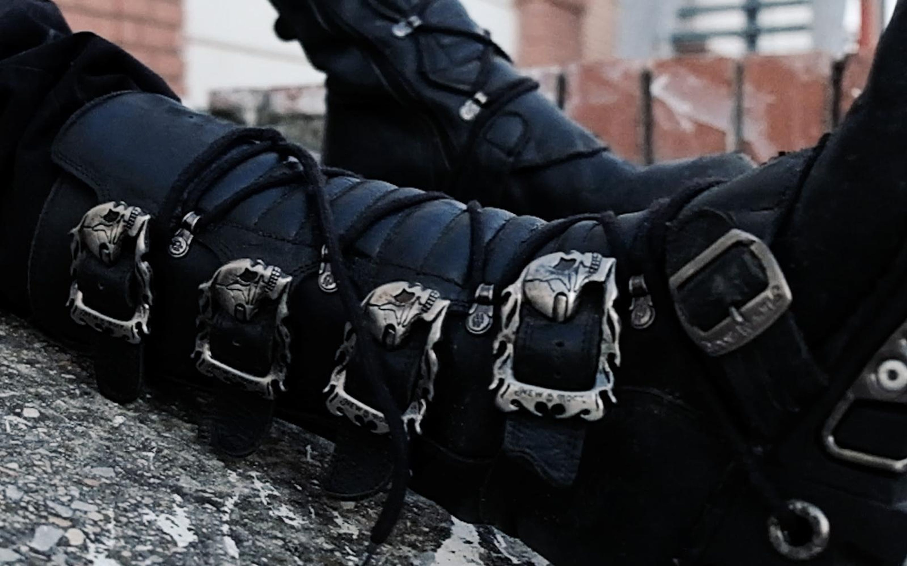 Row of skull-shaped buckles and laces along a black boot resting on rough concrete, brick wall behind