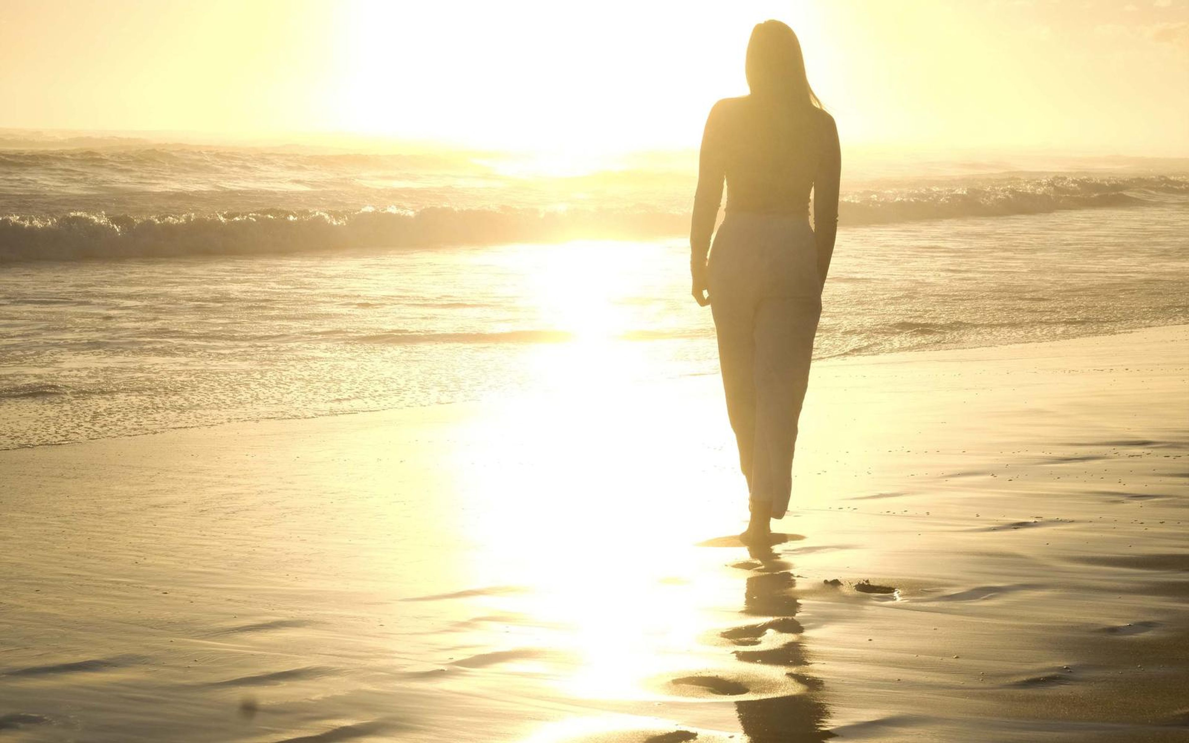 Woman walking away along the water's edge in golden light, wet sand reflecting the sky