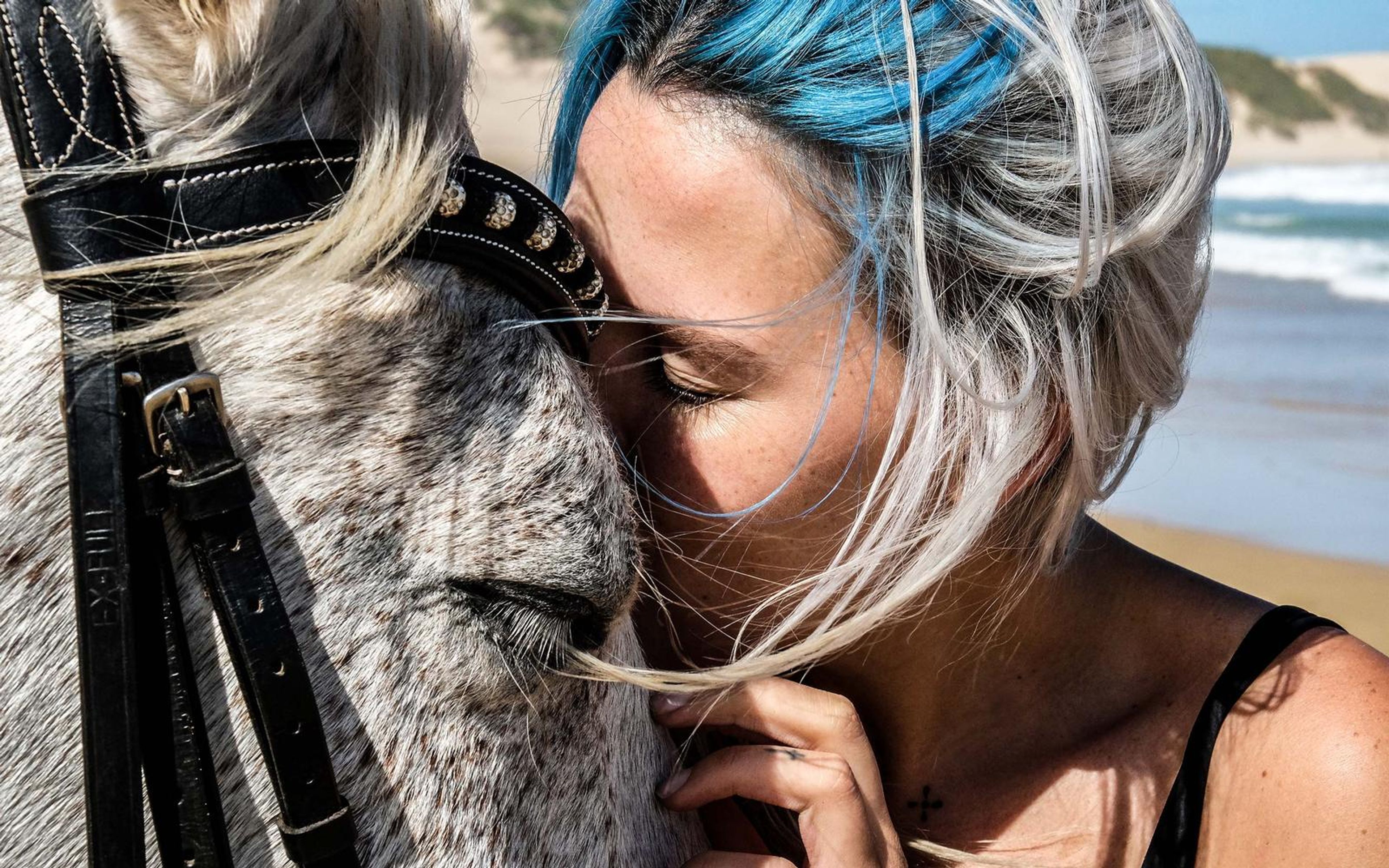Close portrait of a woman with blue hair resting her face on a white horse's cheek, hand near her chin