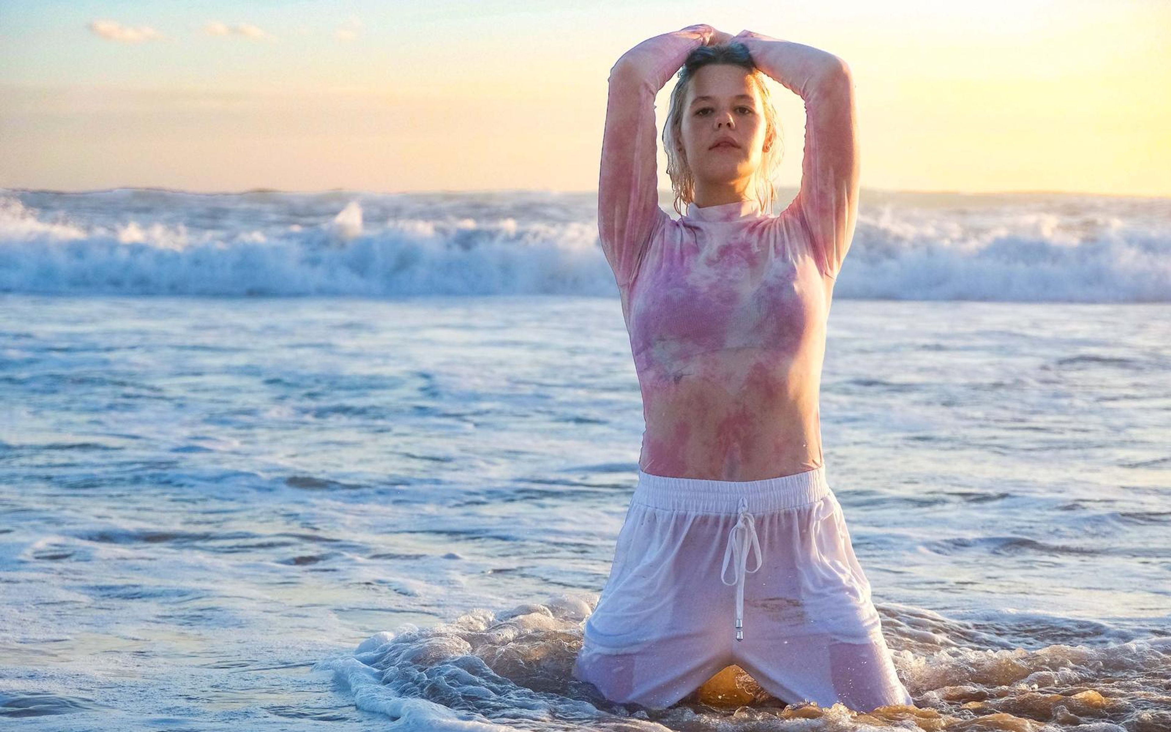 Woman kneeling in shallow surf with arms overhead, wet pink top and white drawstring trousers, bright cloud-lit sky