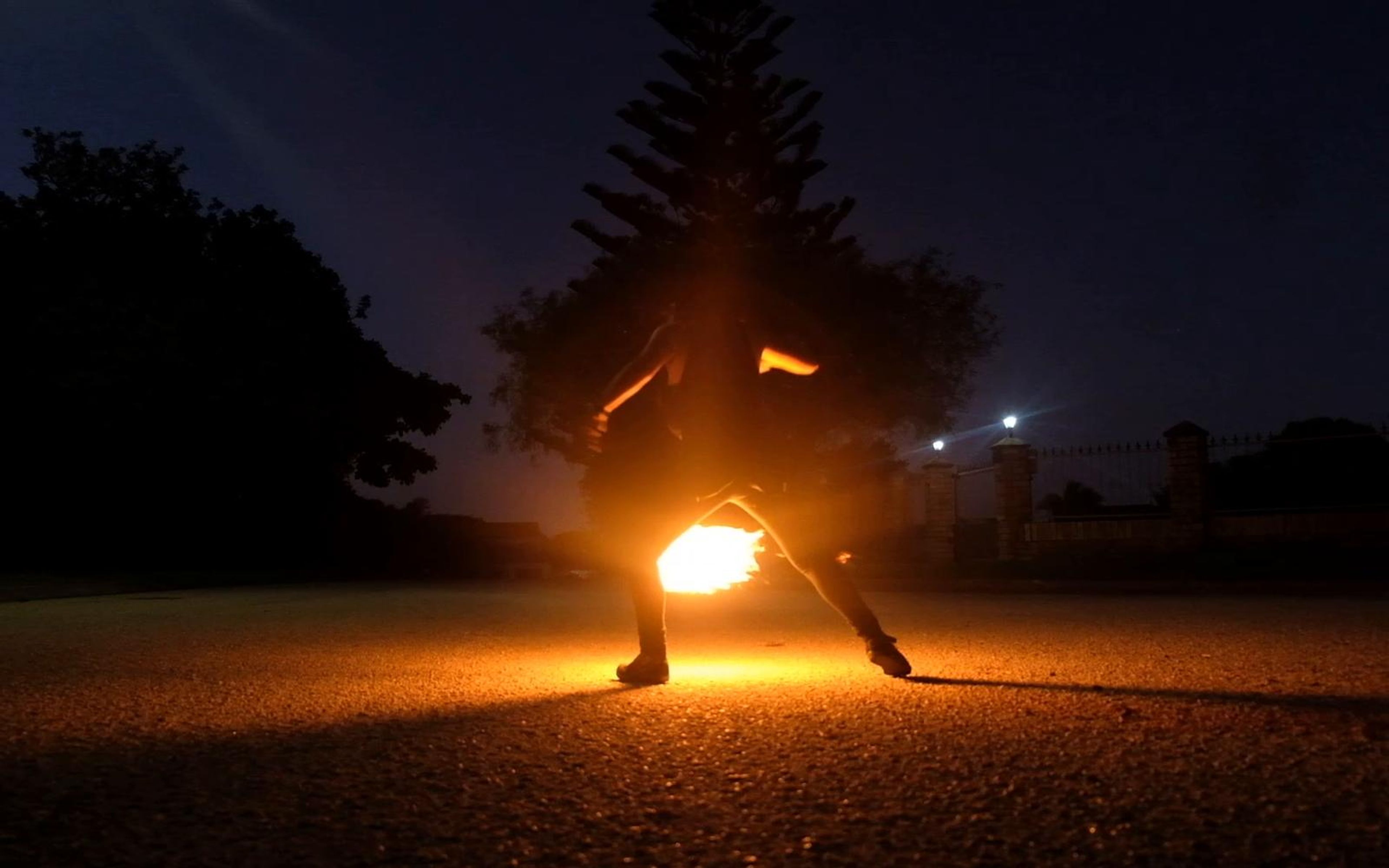 Performer spinning fire in an open lot with tall pines and street lights behind