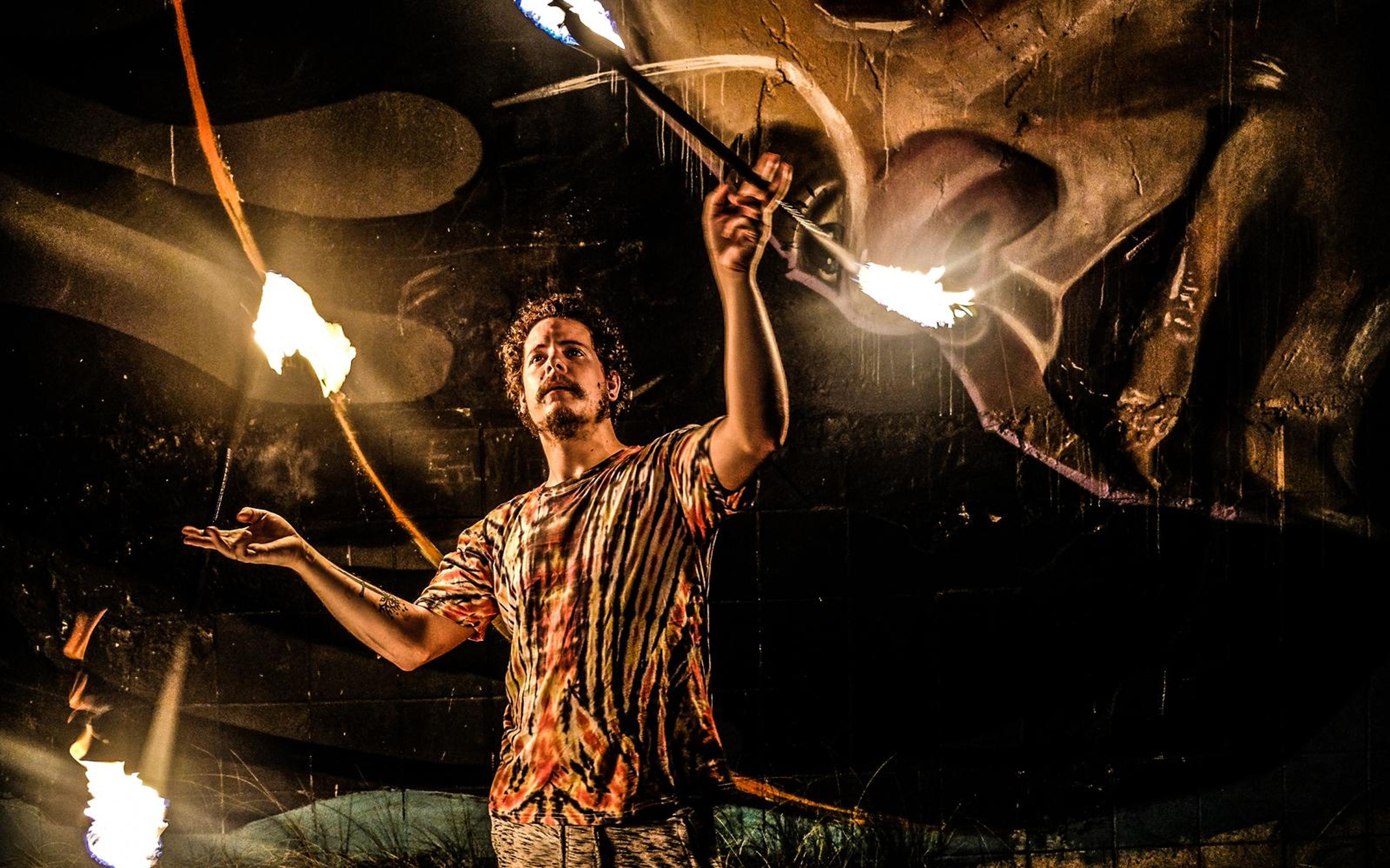 Man in a tie-dye shirt holding two crossed fire staffs in front of a dark painted wall