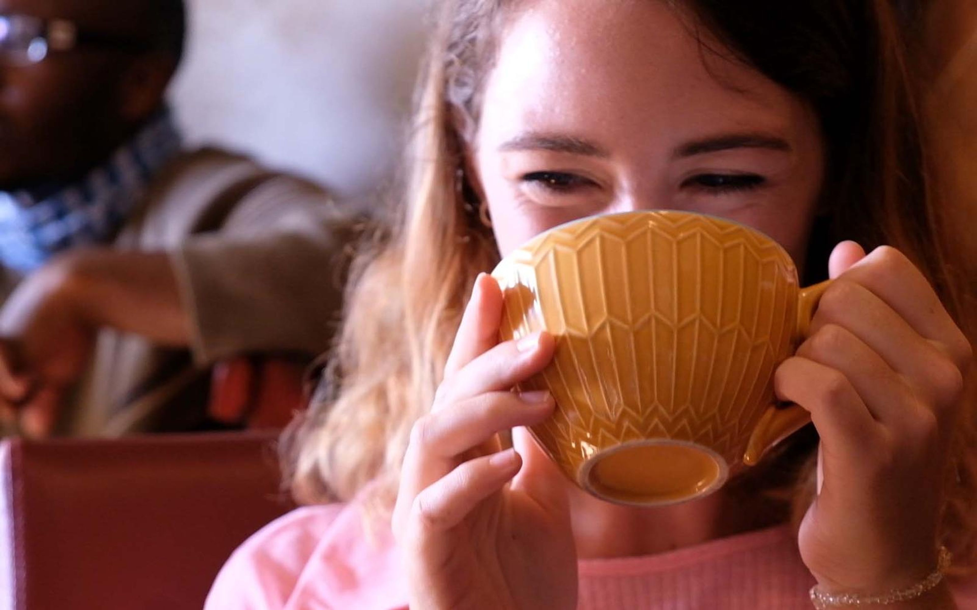 Young woman with eyes closed sipping from a mustard-yellow ceramic cup in a cafe