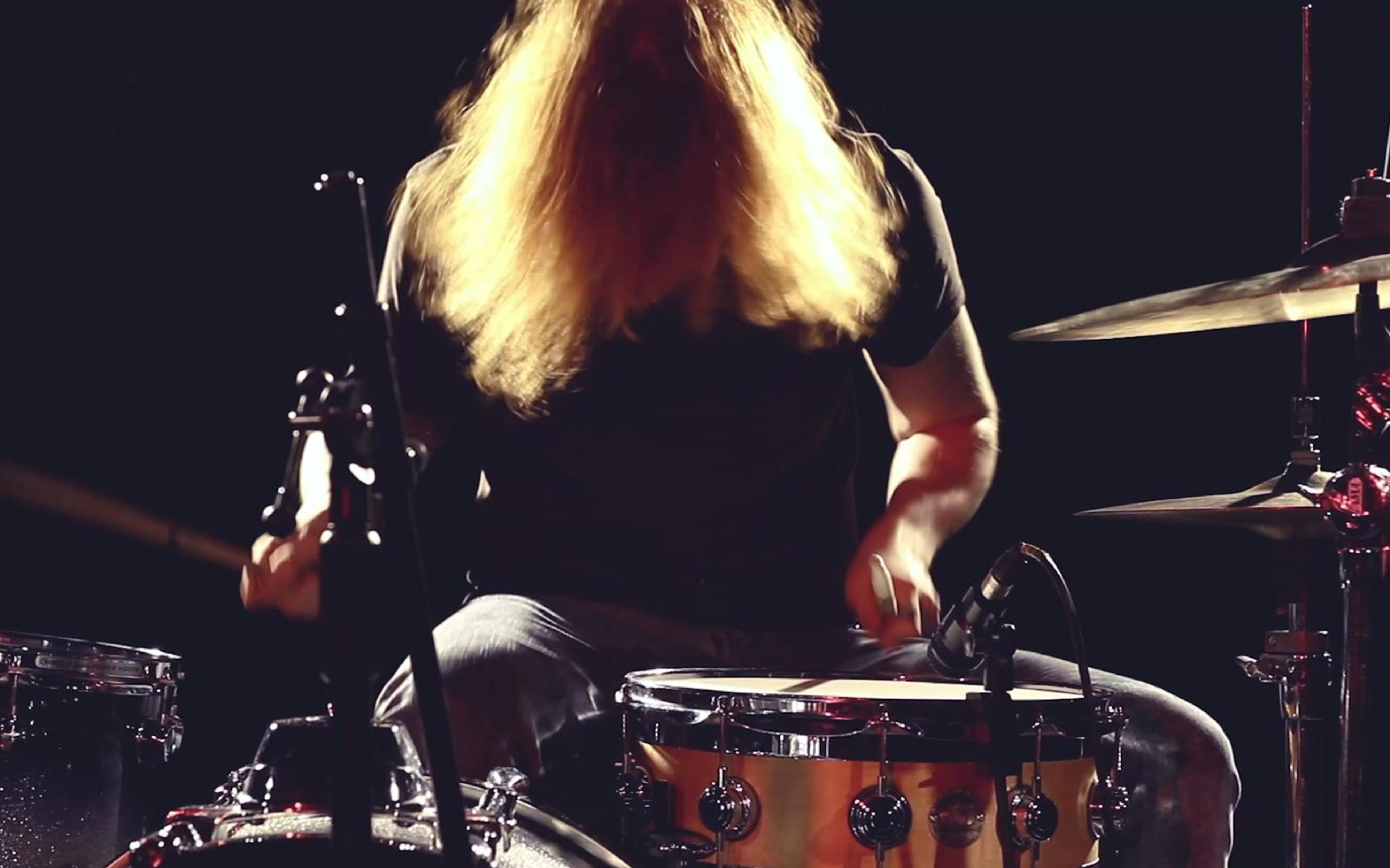 Drummer with long blonde hair backlit in gold light behind a kit and cymbals
