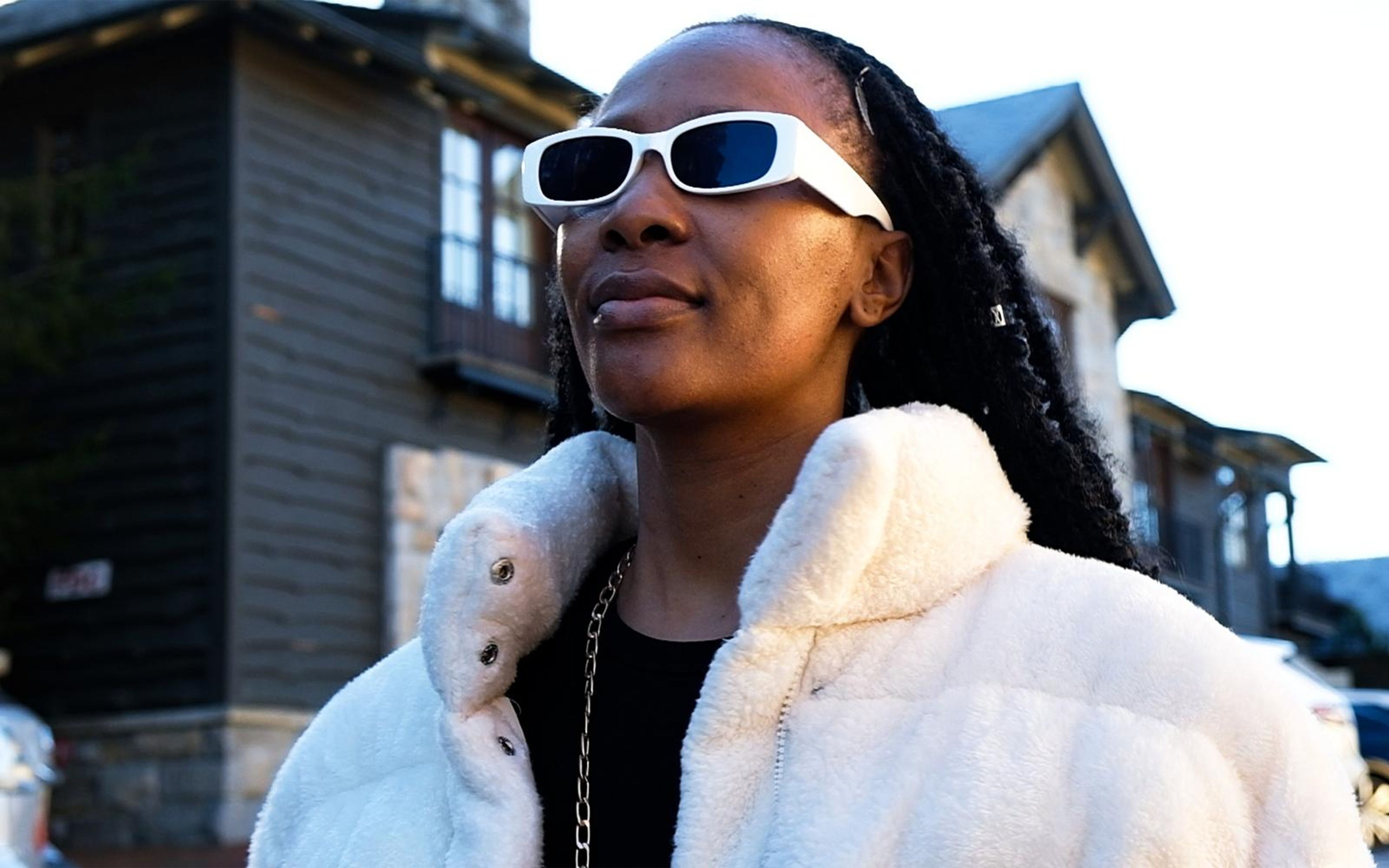 Model in white sunglasses and fur collar looking away, wooden building behind, black top with chain necklace