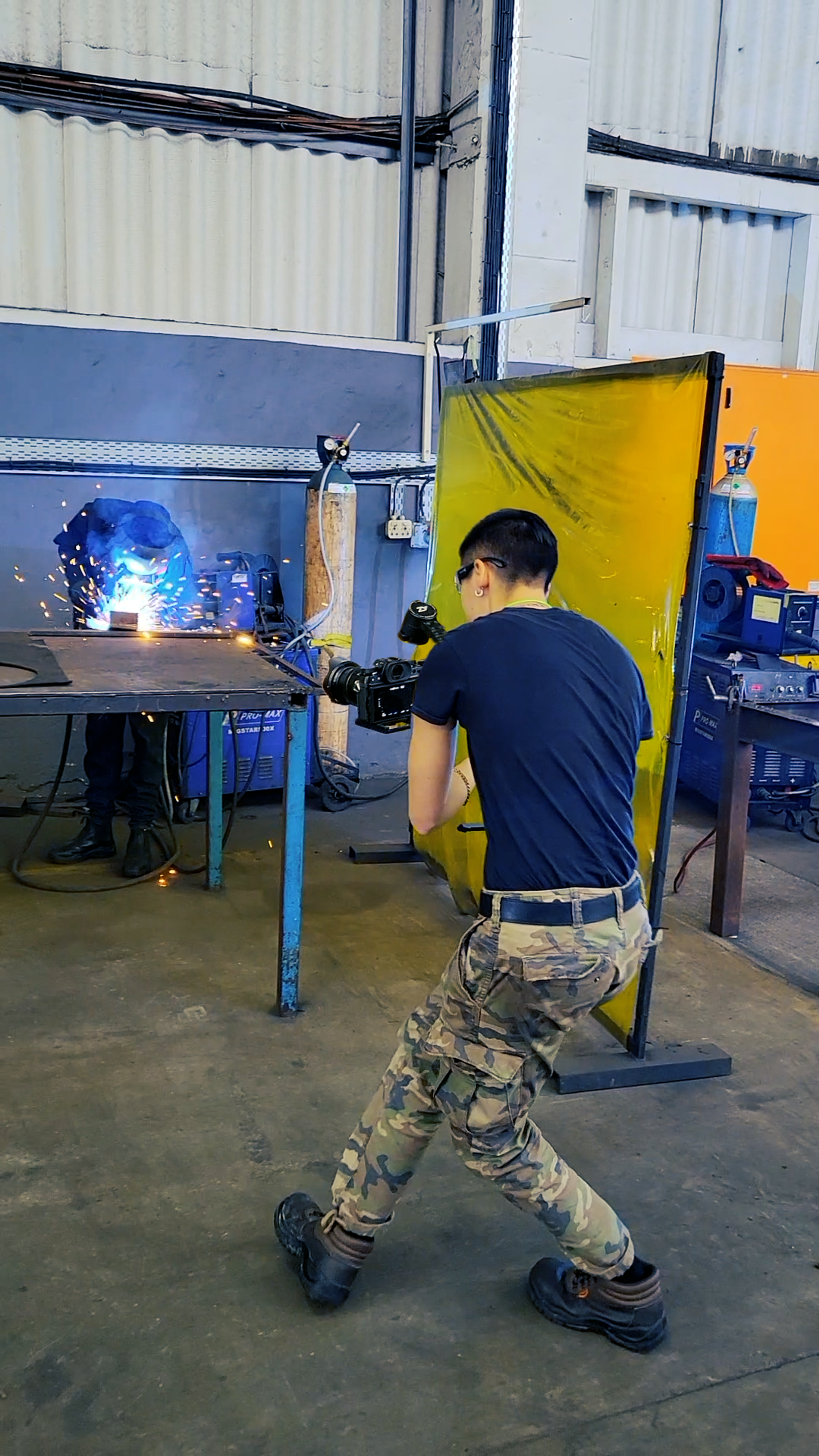 Behind the scenes in a workshop, a camera operator crouching to film a welder at work