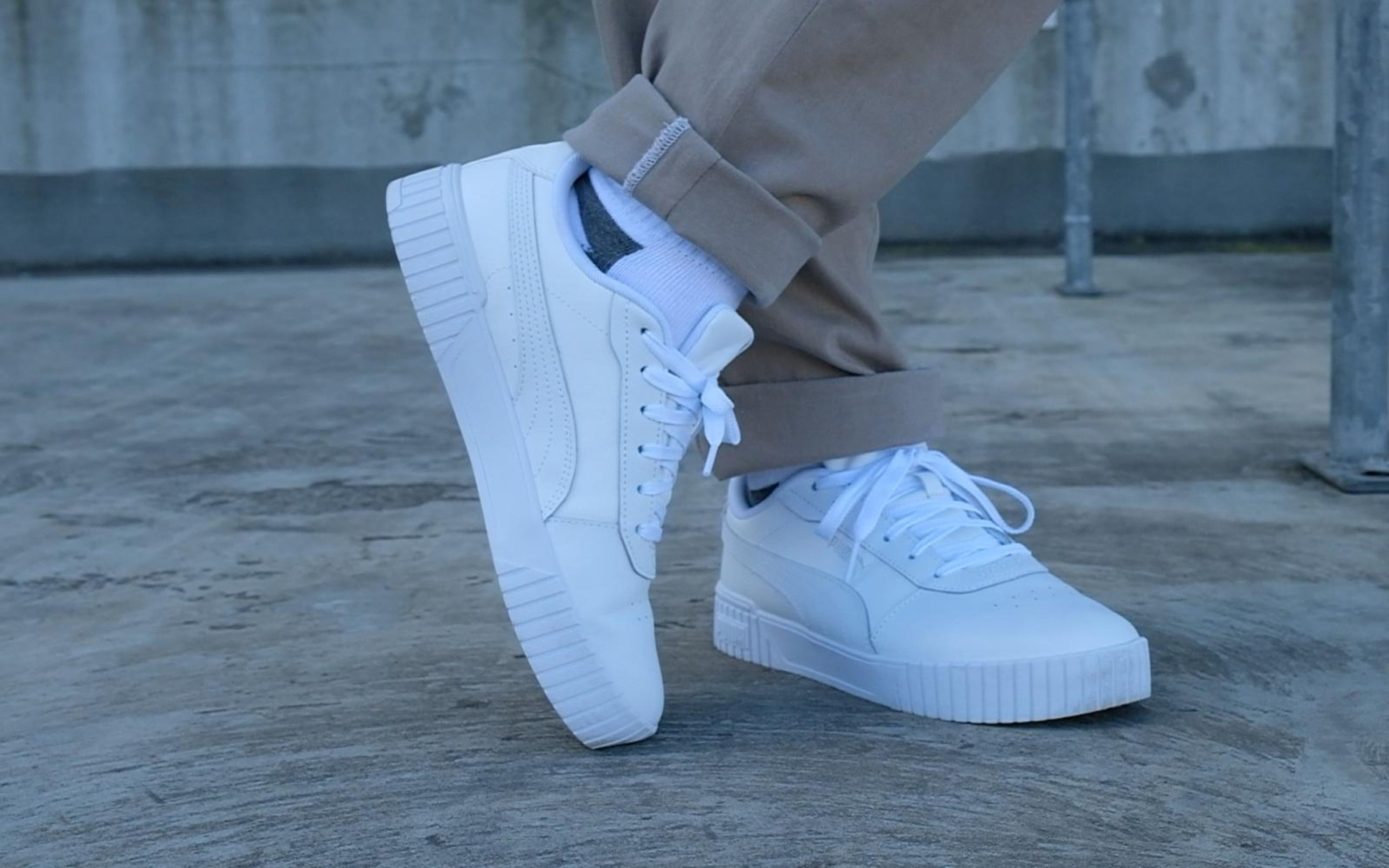 White sneakers with one foot angled on its edge, white sock, cuffed trousers, concrete wall behind