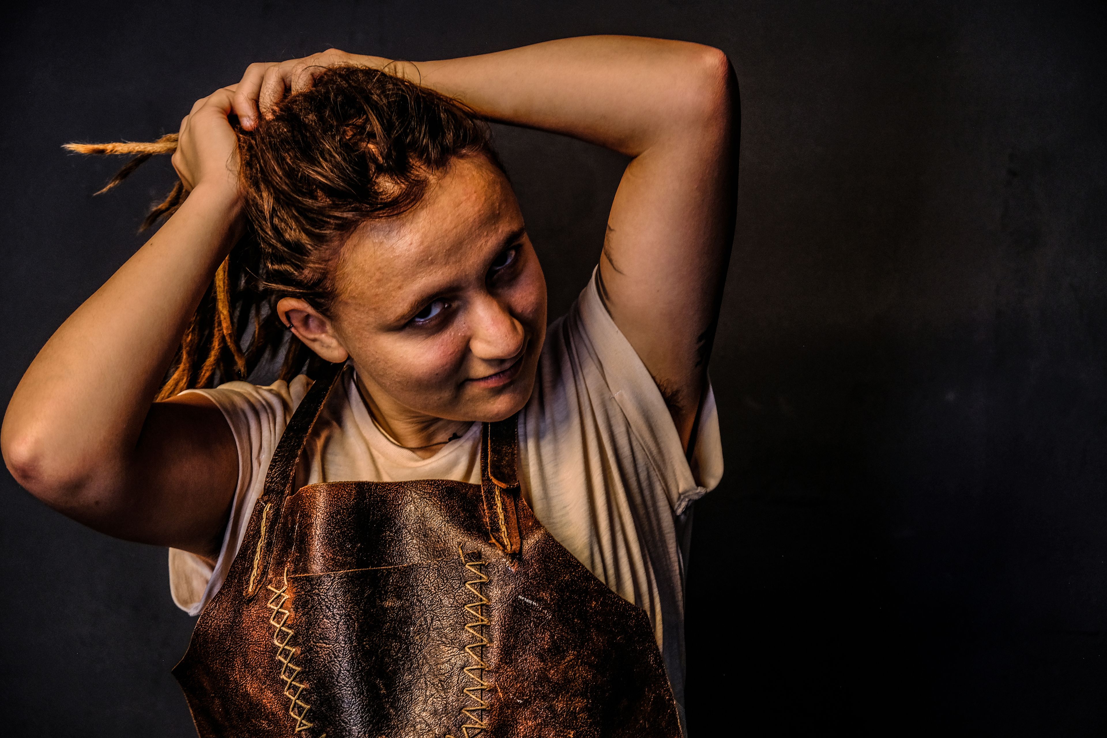 Warm-toned portrait, subject gathering dreadlocks up with both hands, wearing a leather apron