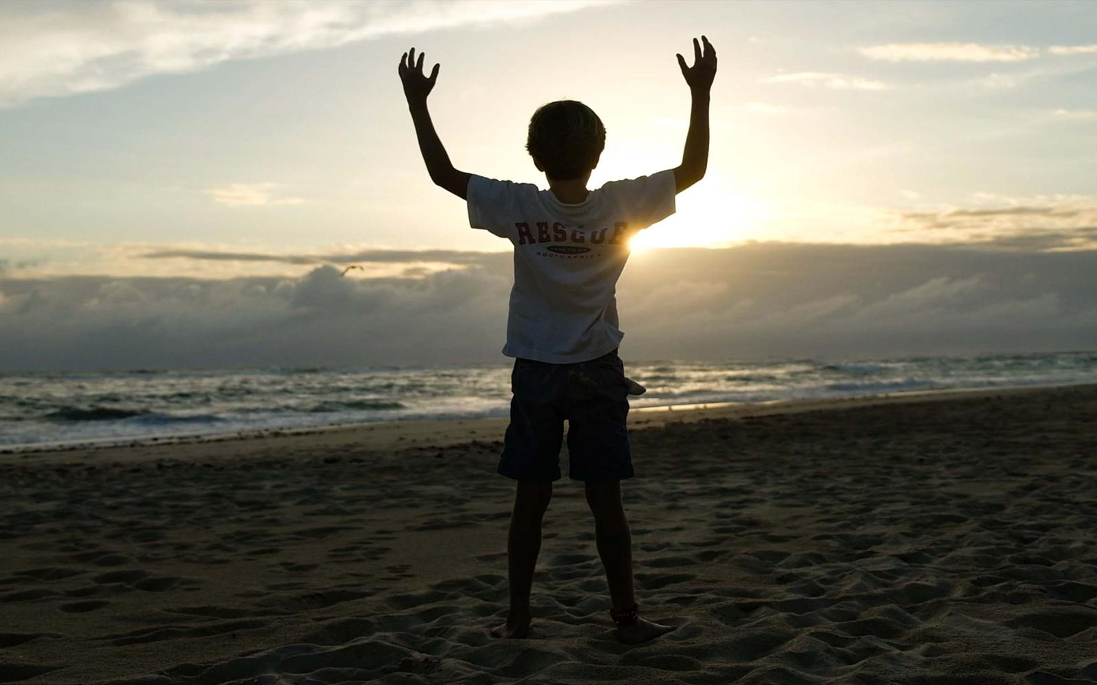 Silhouetted boy with raised arms on a beach at sunset with a cloud bank on the horizon
