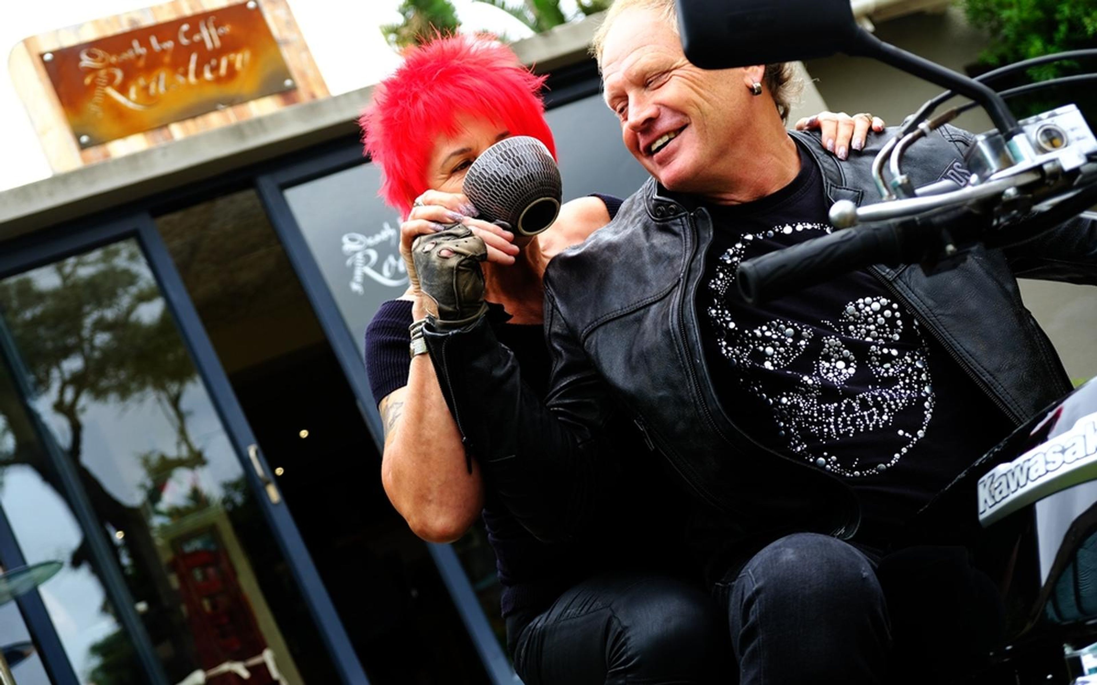 Woman with red hair sipping from a cup while the man beside her smiles, seated on a motorbike outside a cafe