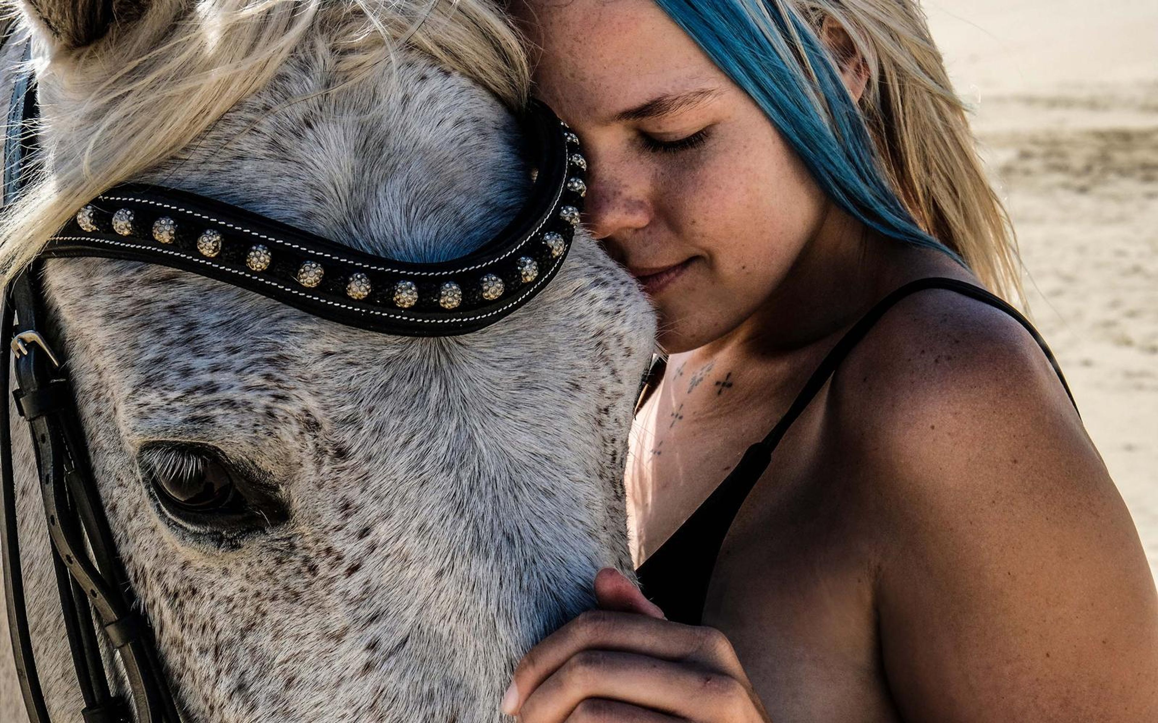 Close portrait of a woman with blue-streaked hair and bare shoulders, eyes closed, pressed against a studded-bridle horse face