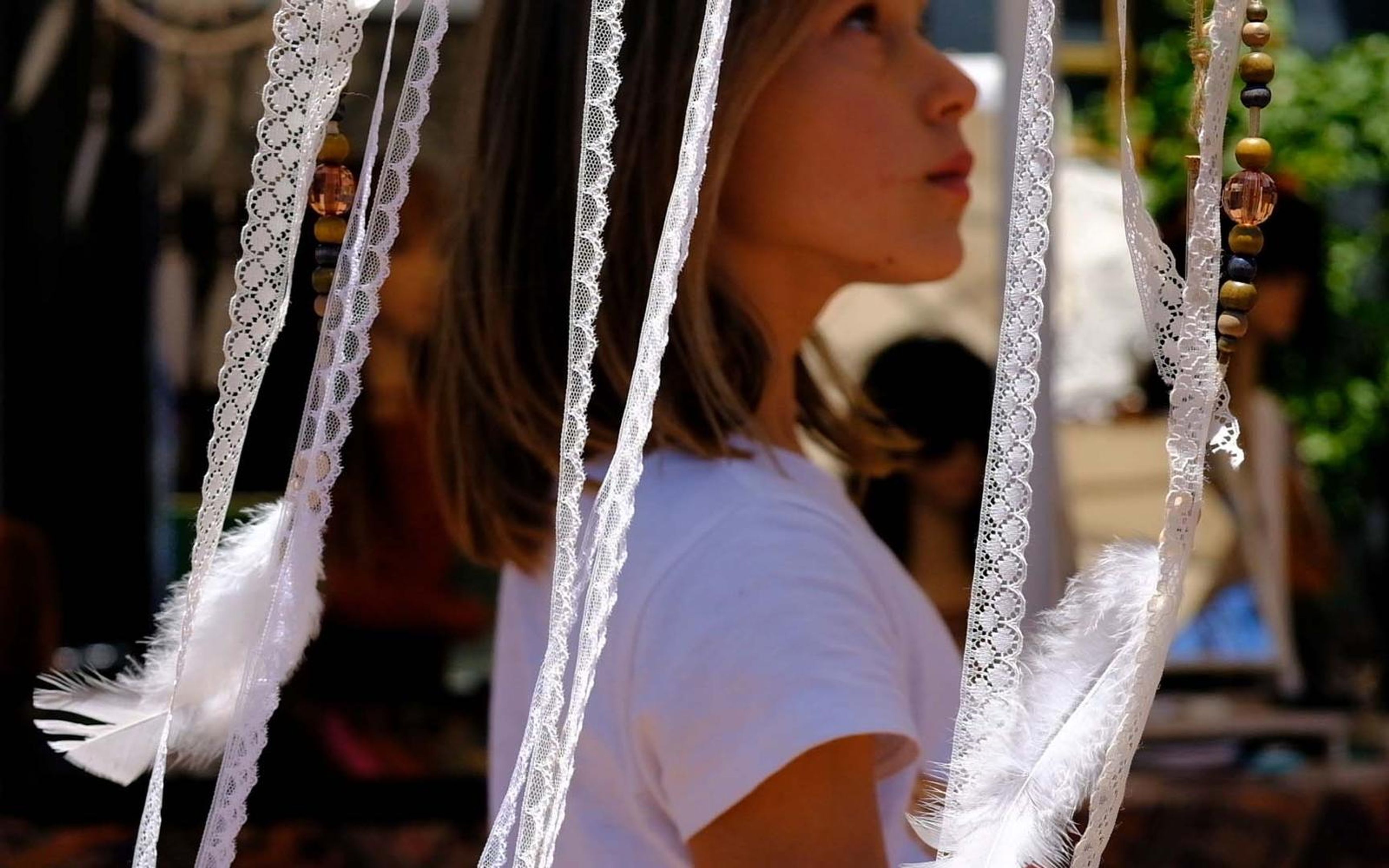 Girl in a white top looking off to the side behind hanging lace and feather charms at a market stall