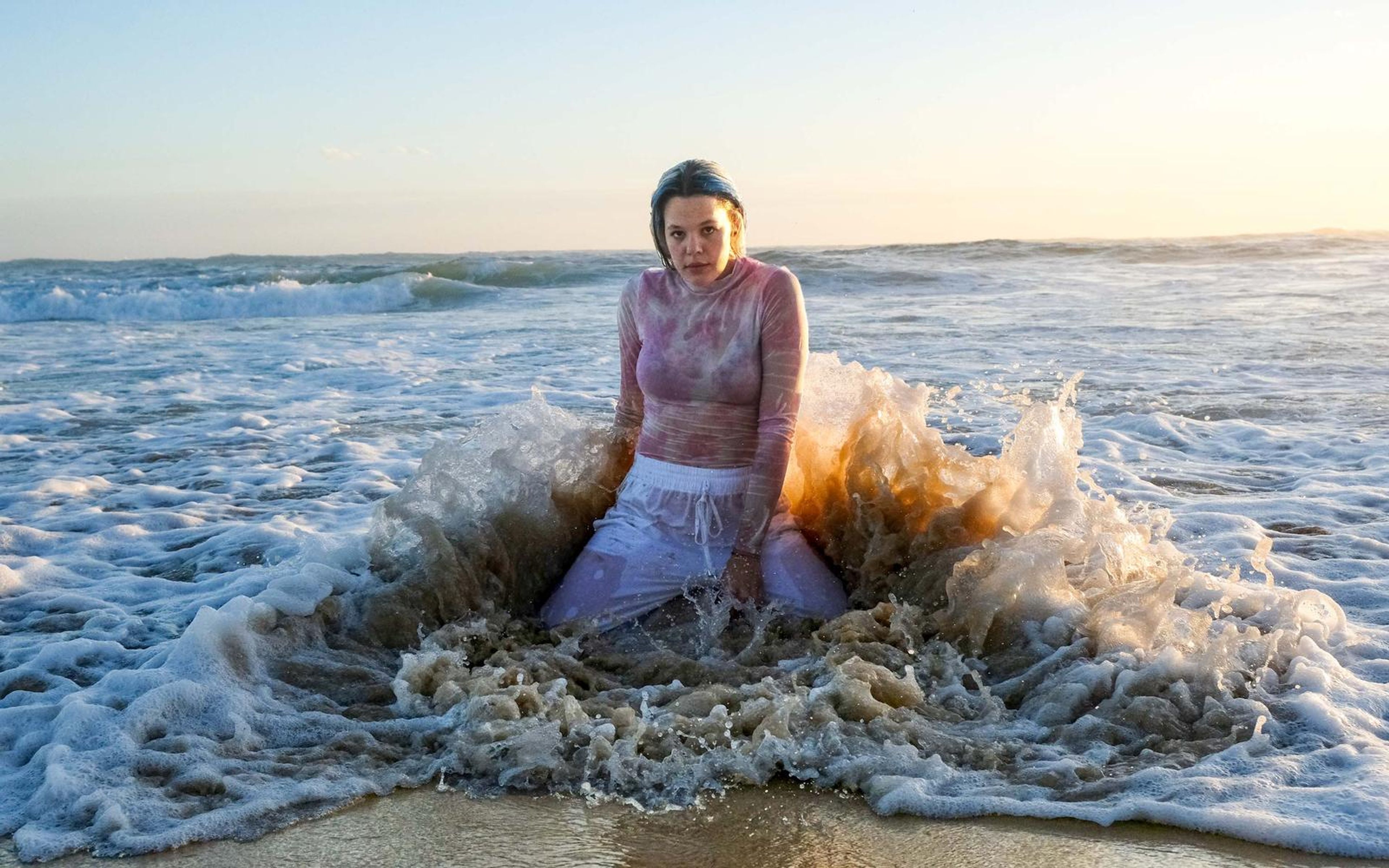 Woman sitting in the surf as a wave crashes against her, amber spray lit by the low sun, pale sky above
