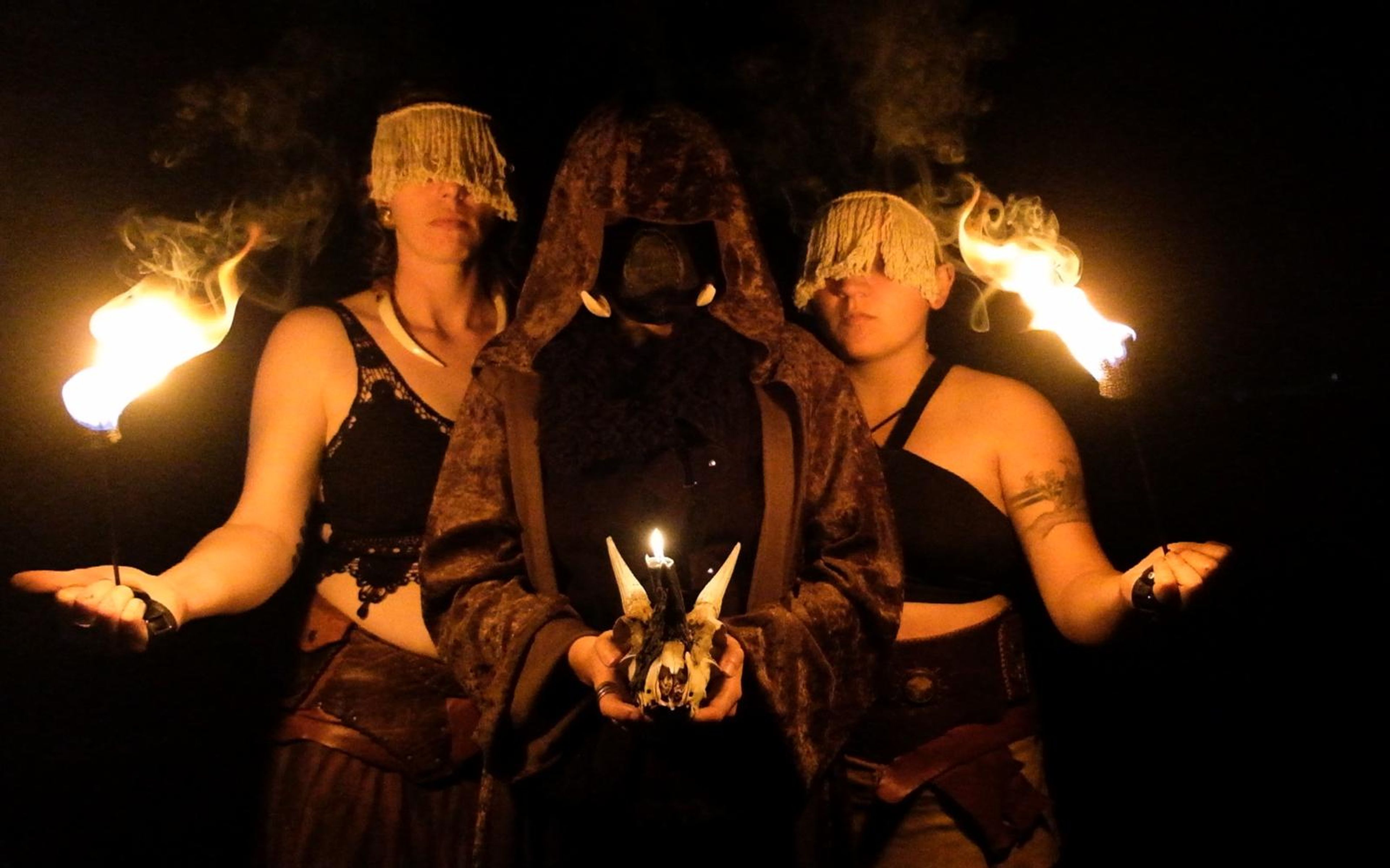 Three costumed performers in a dark clearing, two with fringed head coverings, a hooded figure holding a horned skull