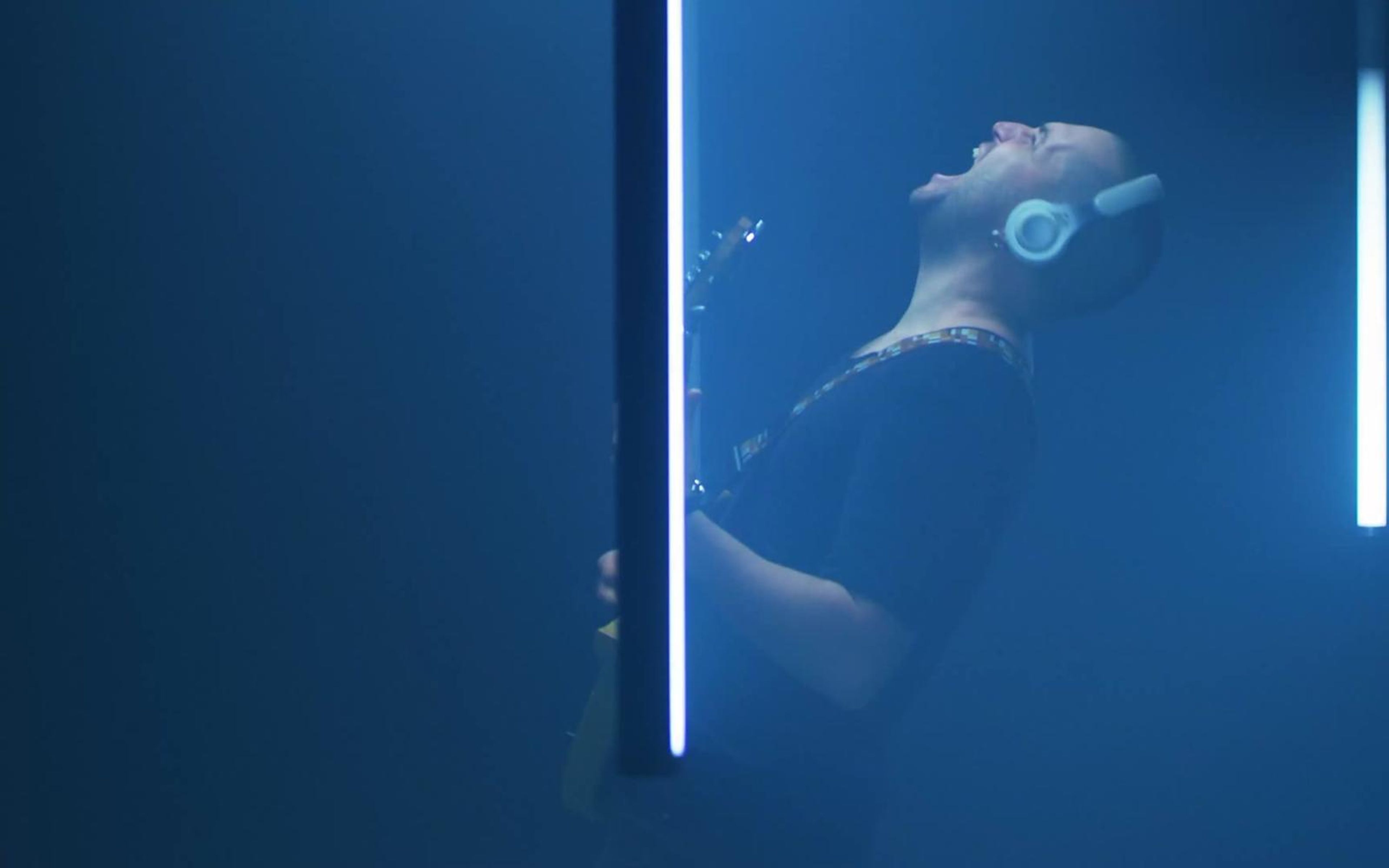 Bald guitarist in headphones leaning back mid-song, lit in blue with a bright light tube beside him