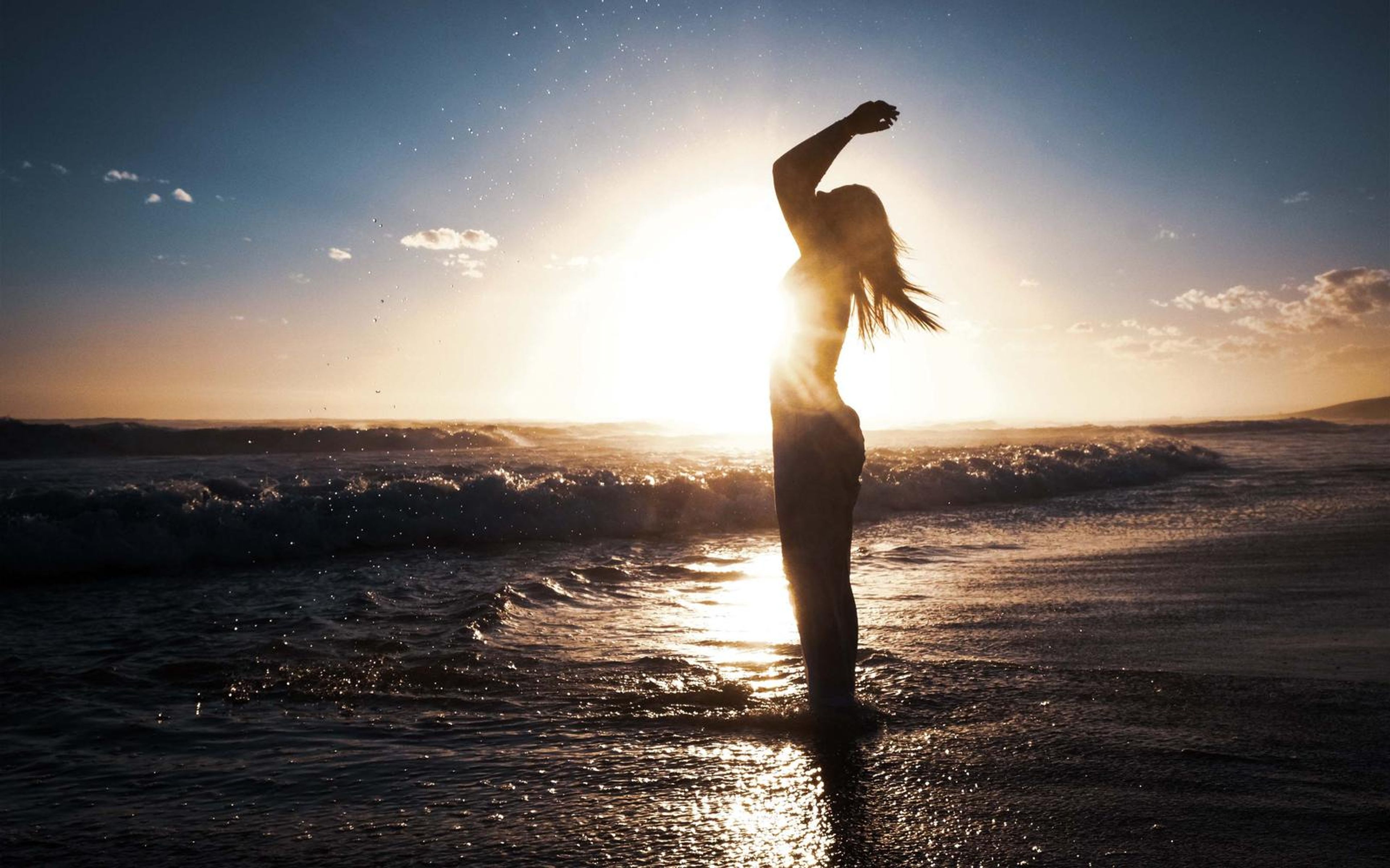 Backlit woman with one arm raised against a deep sunset sky over the shoreline