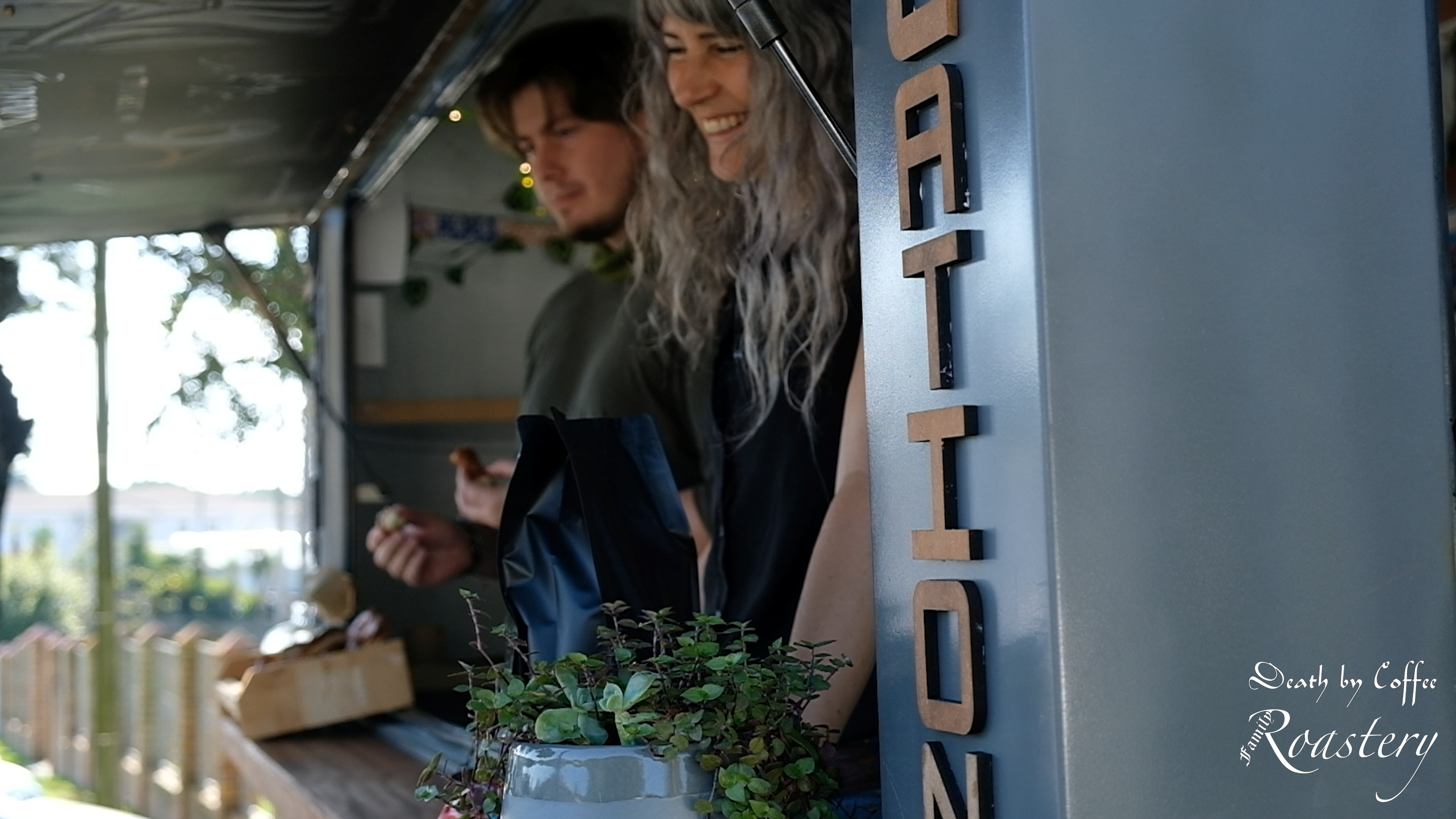 Two baristas smiling and serving from the window of Death By Coffee's mobile coffee trailer, greenery and wood accents on the counter
