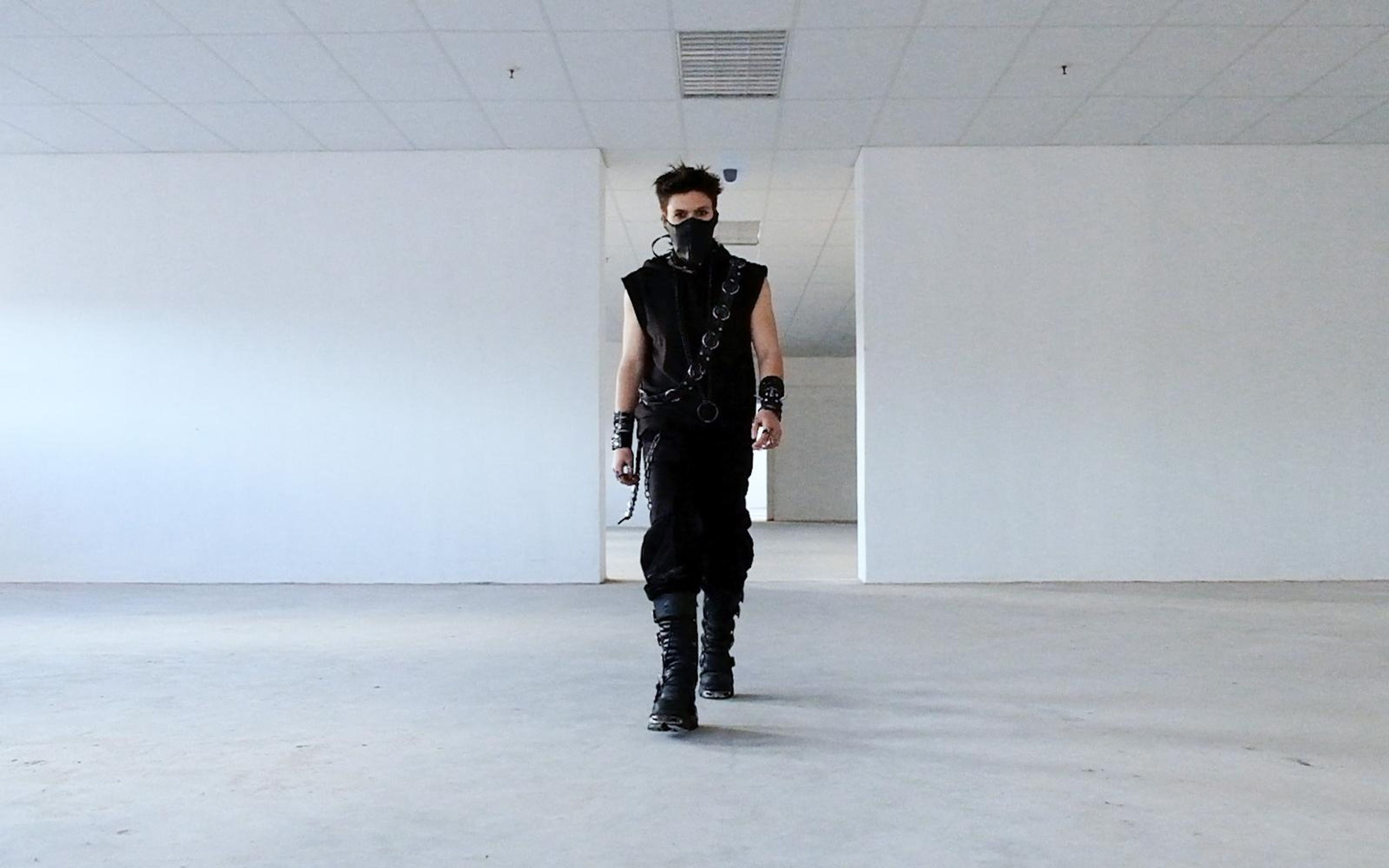 Masked figure in black and New Rock boots walking straight toward camera down the middle of a white warehouse between two walls
