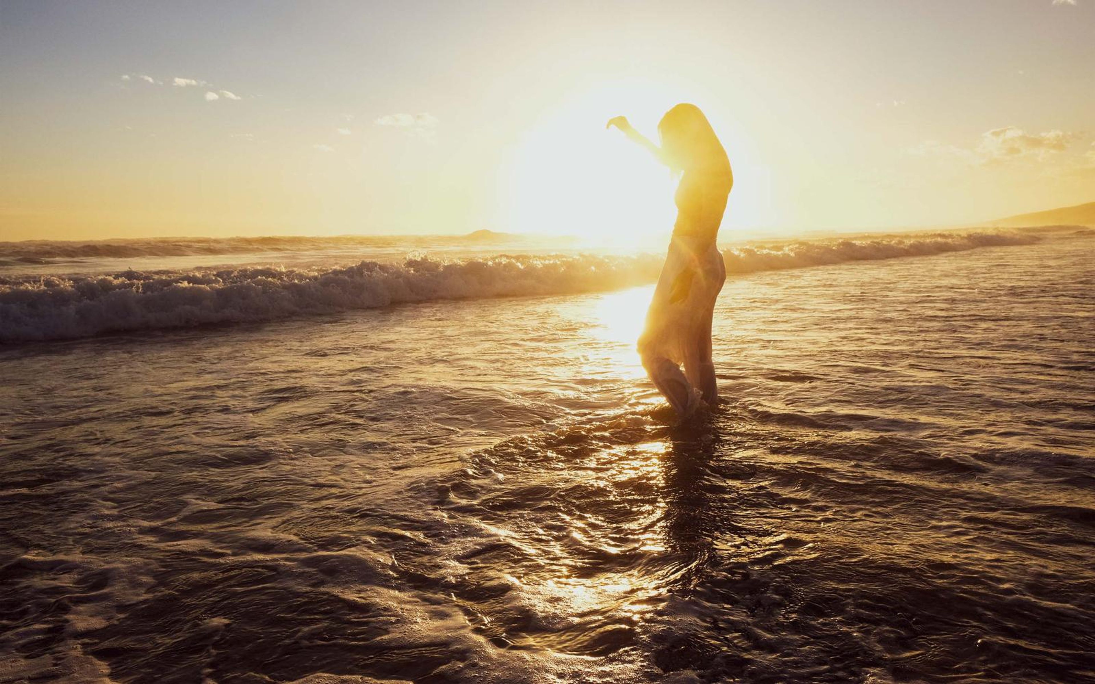 Woman standing in the surf holding one arm out to the side, warm light on the water