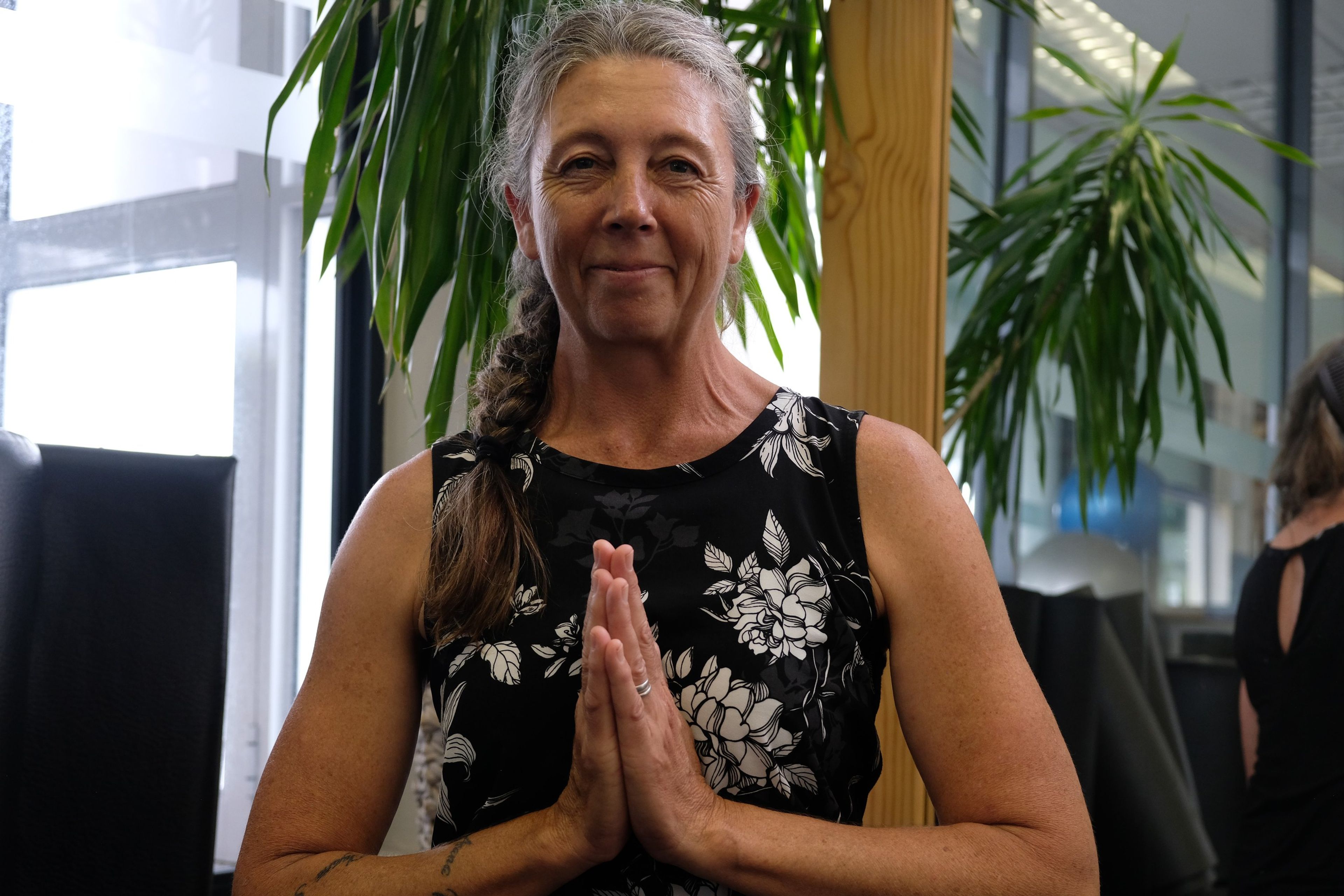 Seated instructor with a long grey braid and floral top, hands in prayer position, smiling
