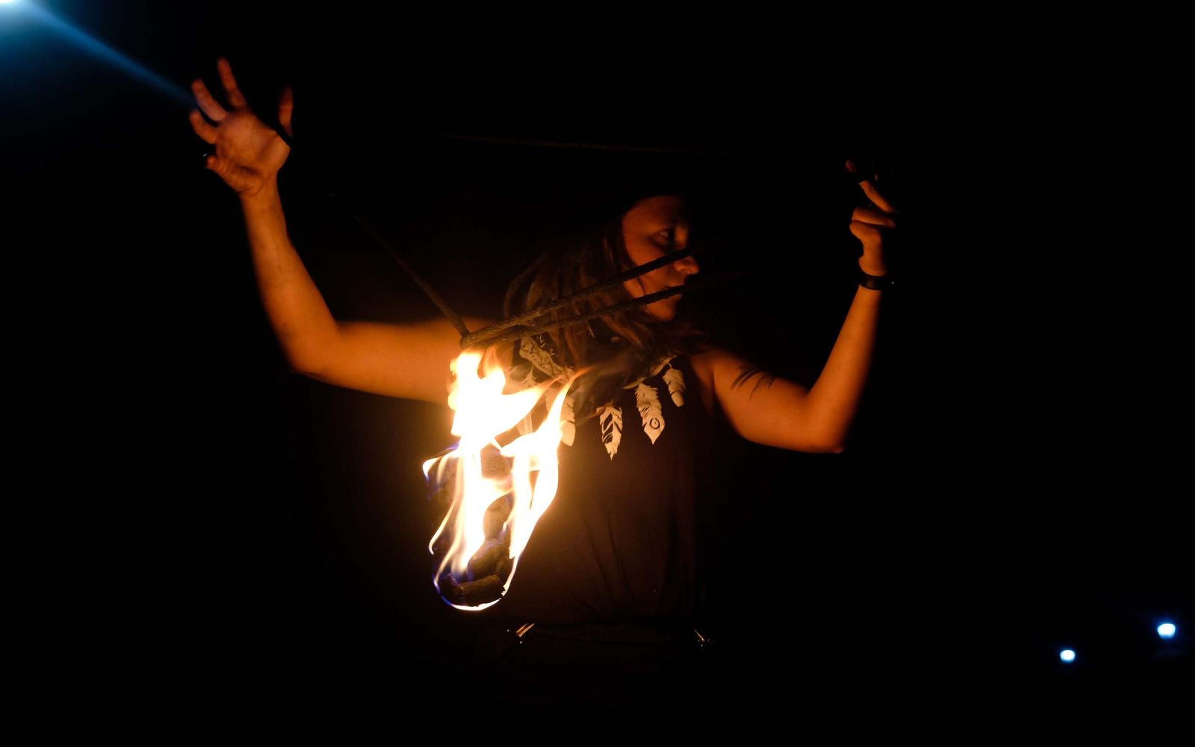 Performer with one hand raised and a flame at the chest, blue lights in the dark at right