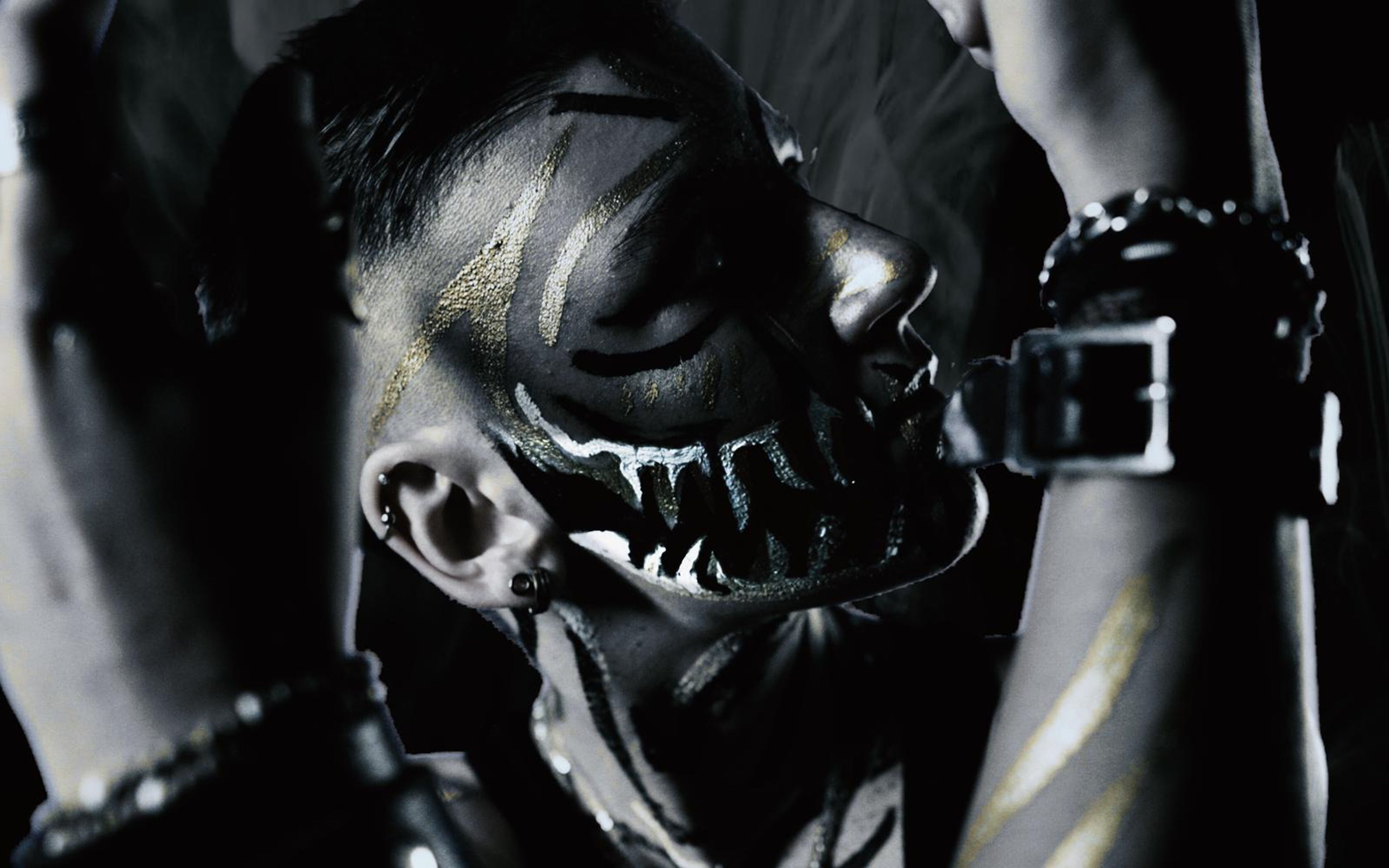 Wide crop of a painted face in profile with white fang markings, a raised hand and a leather wrist cuff in black and white
