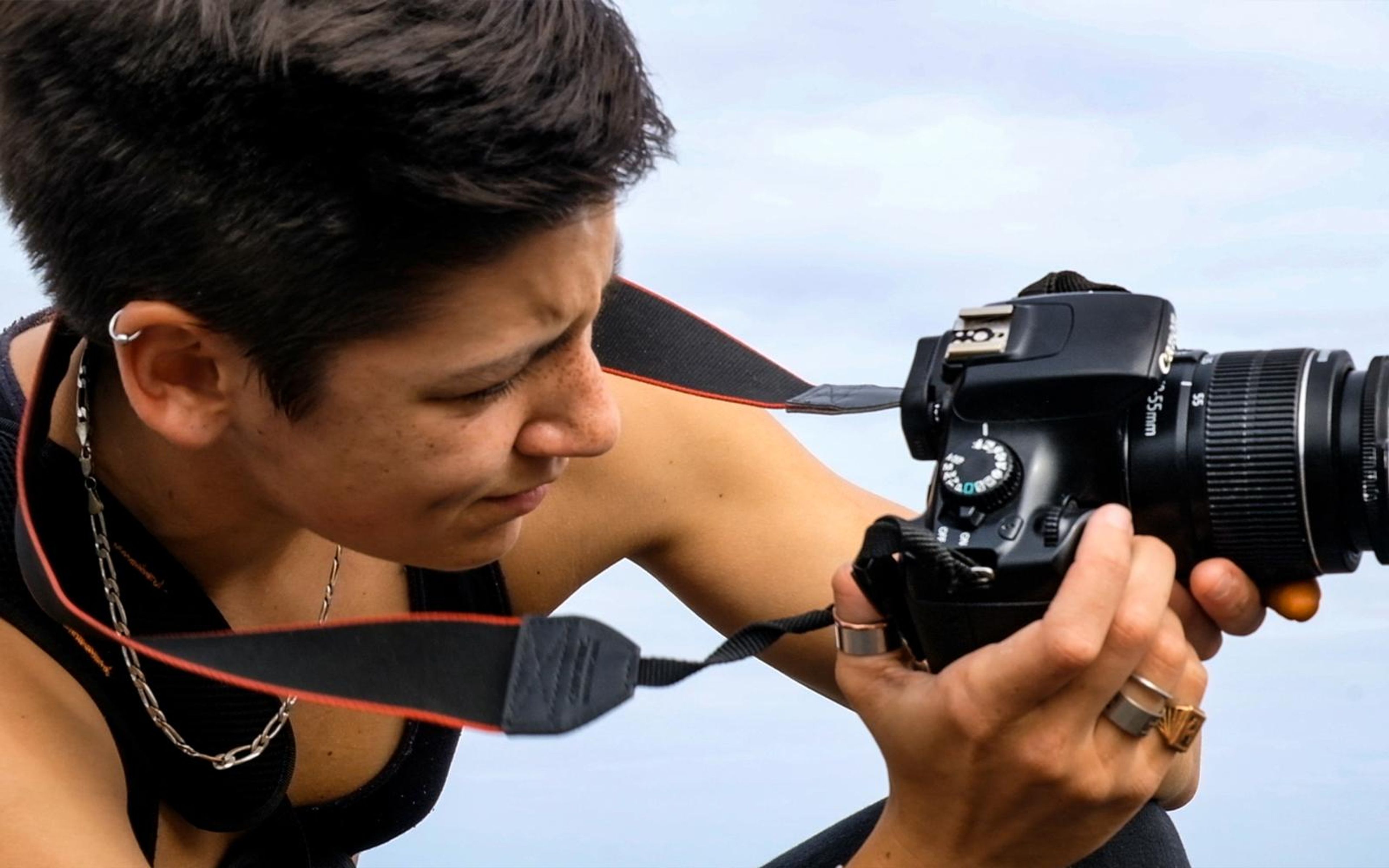 Close view of a woman with short dark hair leaning in to look at her camera against a pale sky