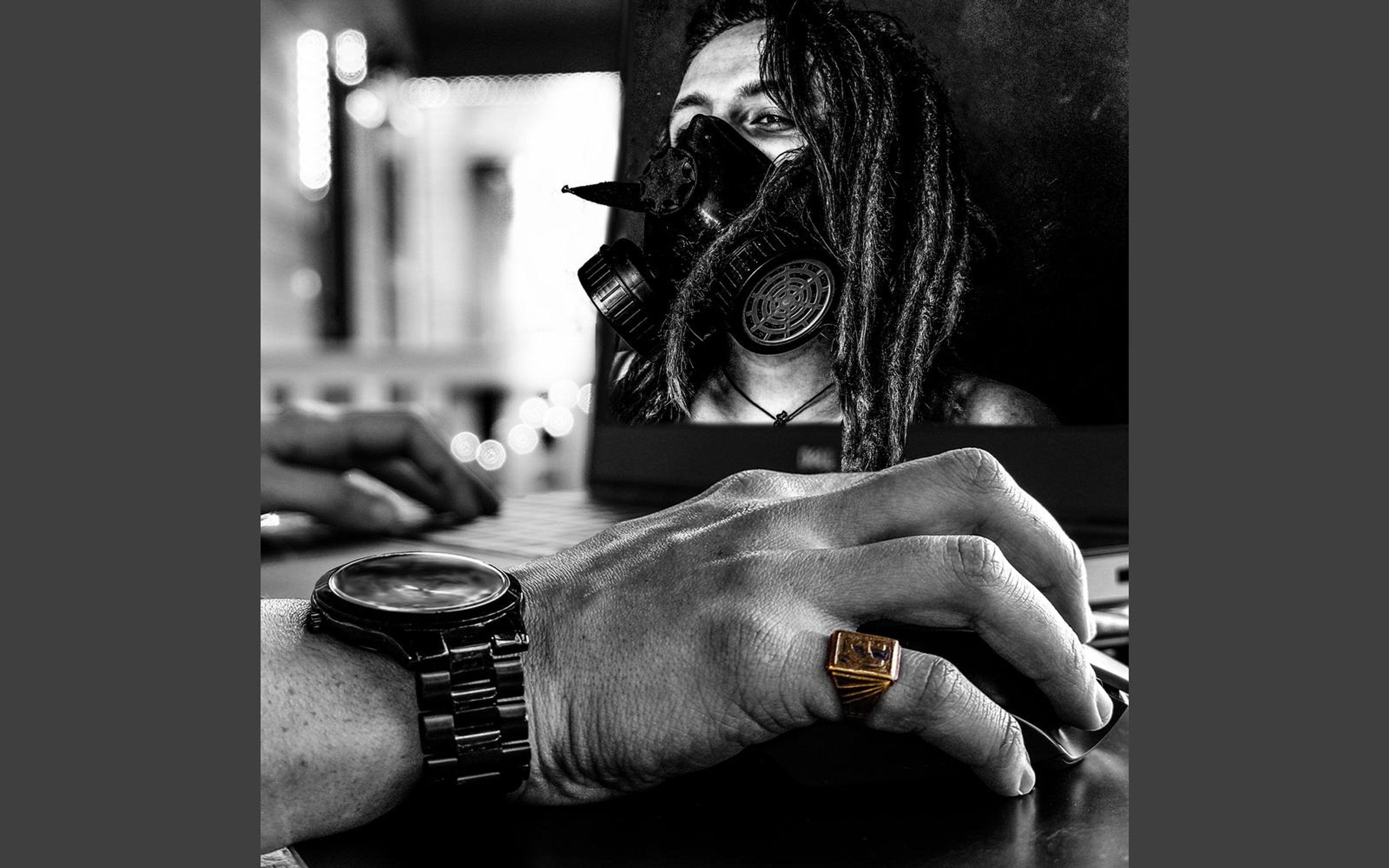 Black and white square composite of a hand on a laptop keyboard with a dreadlocked person in a gas mask rising out of the screen
