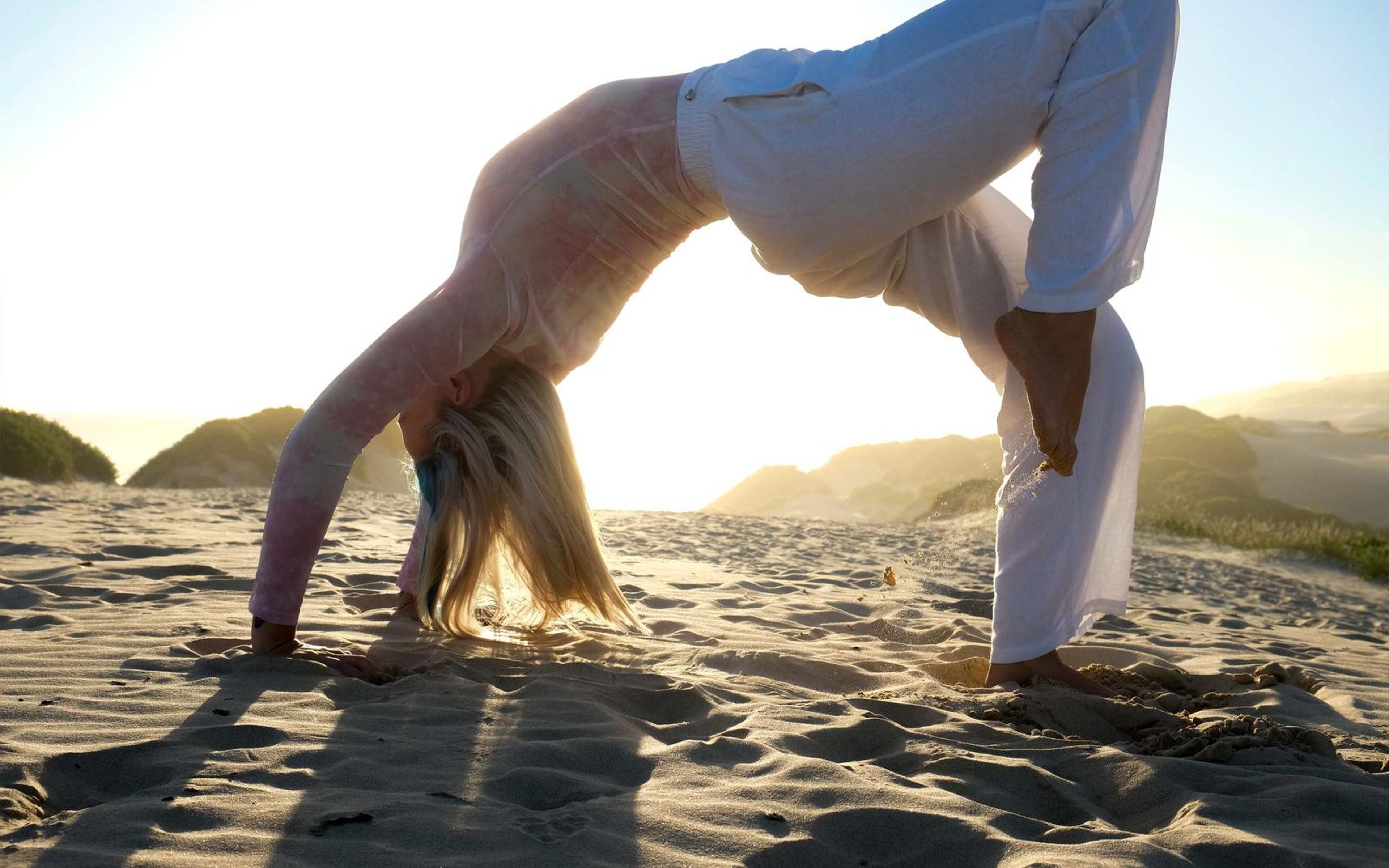 Woman in white trousers and top arching in a backbend on the sand, long shadows