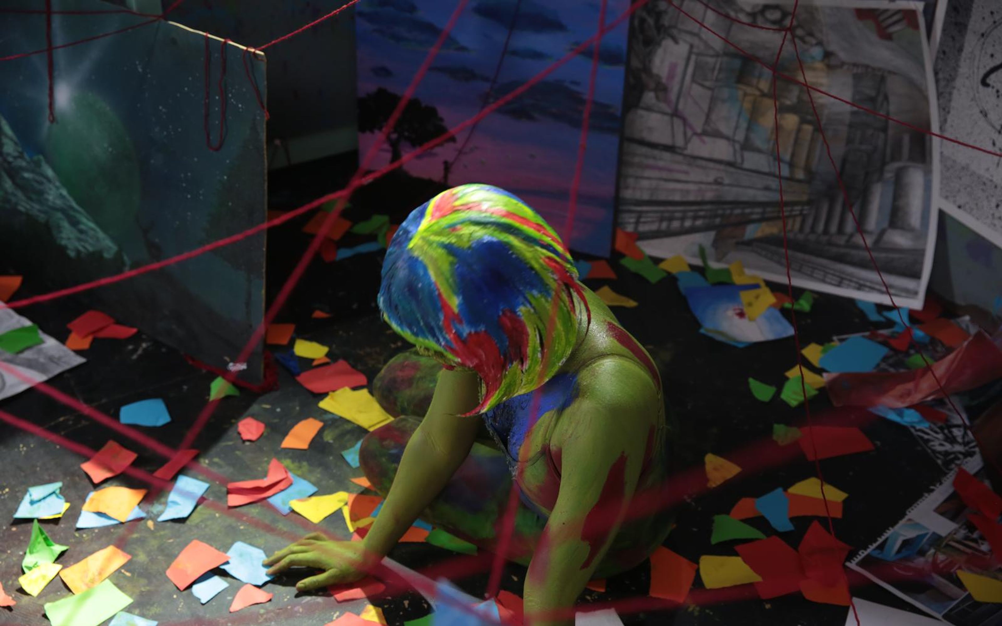 Painted figure crawling across a floor scattered with coloured paper scraps and red thread
