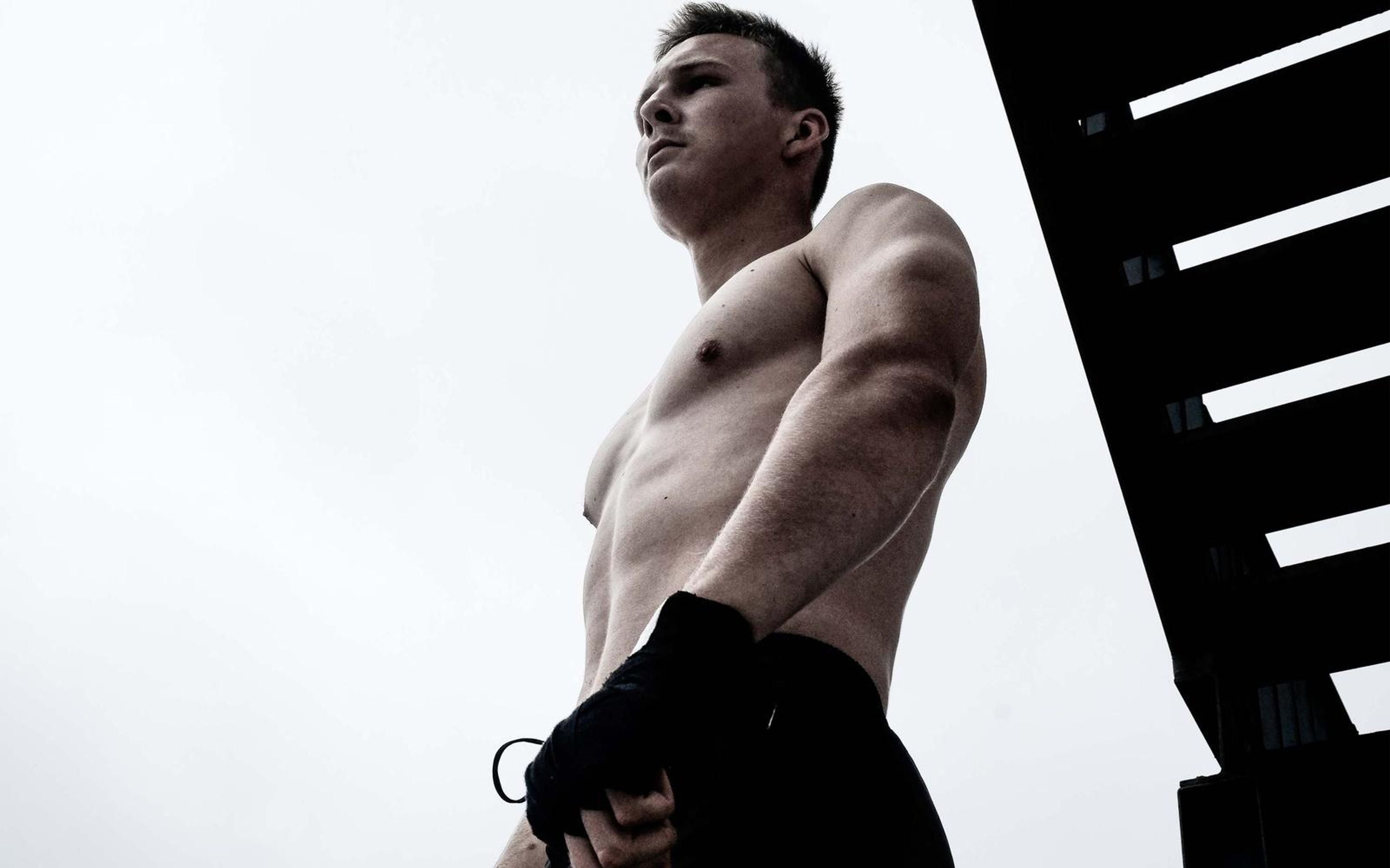 Low angle shot of a man beside a dark metal staircase against a pale sky, wrapped hands, head turned