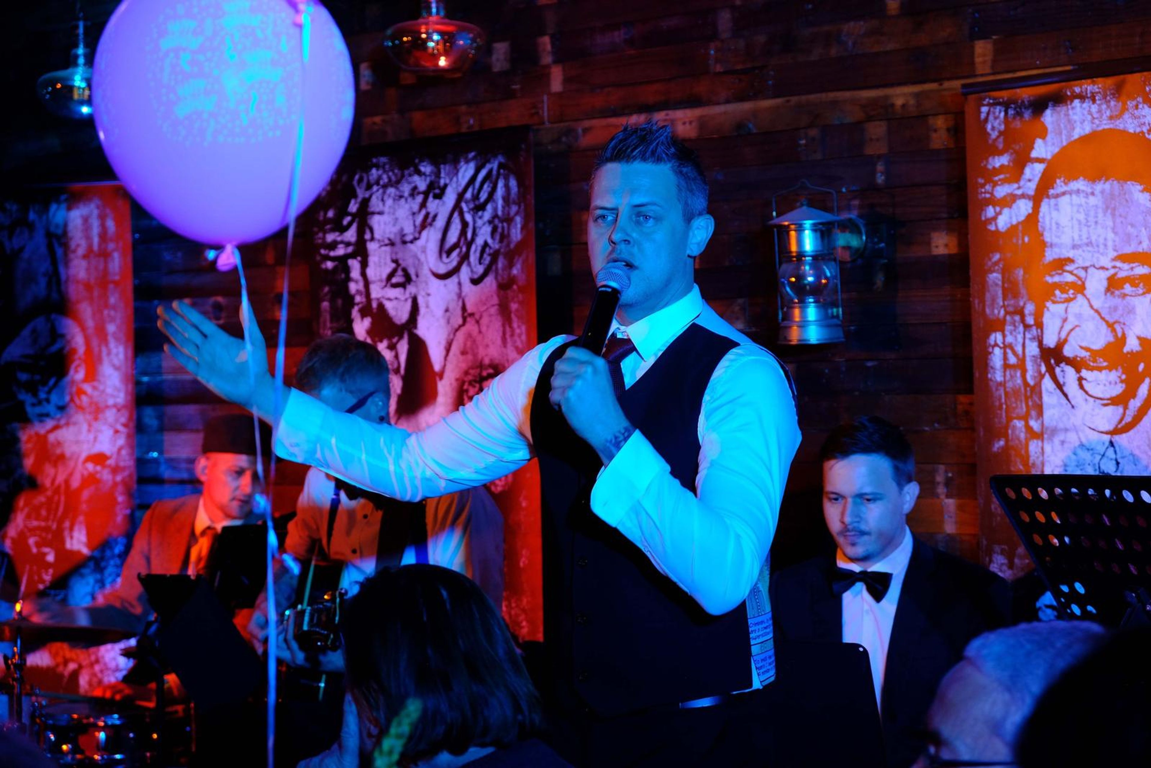 Singer in a waistcoat with his arms out under blue light and a balloon