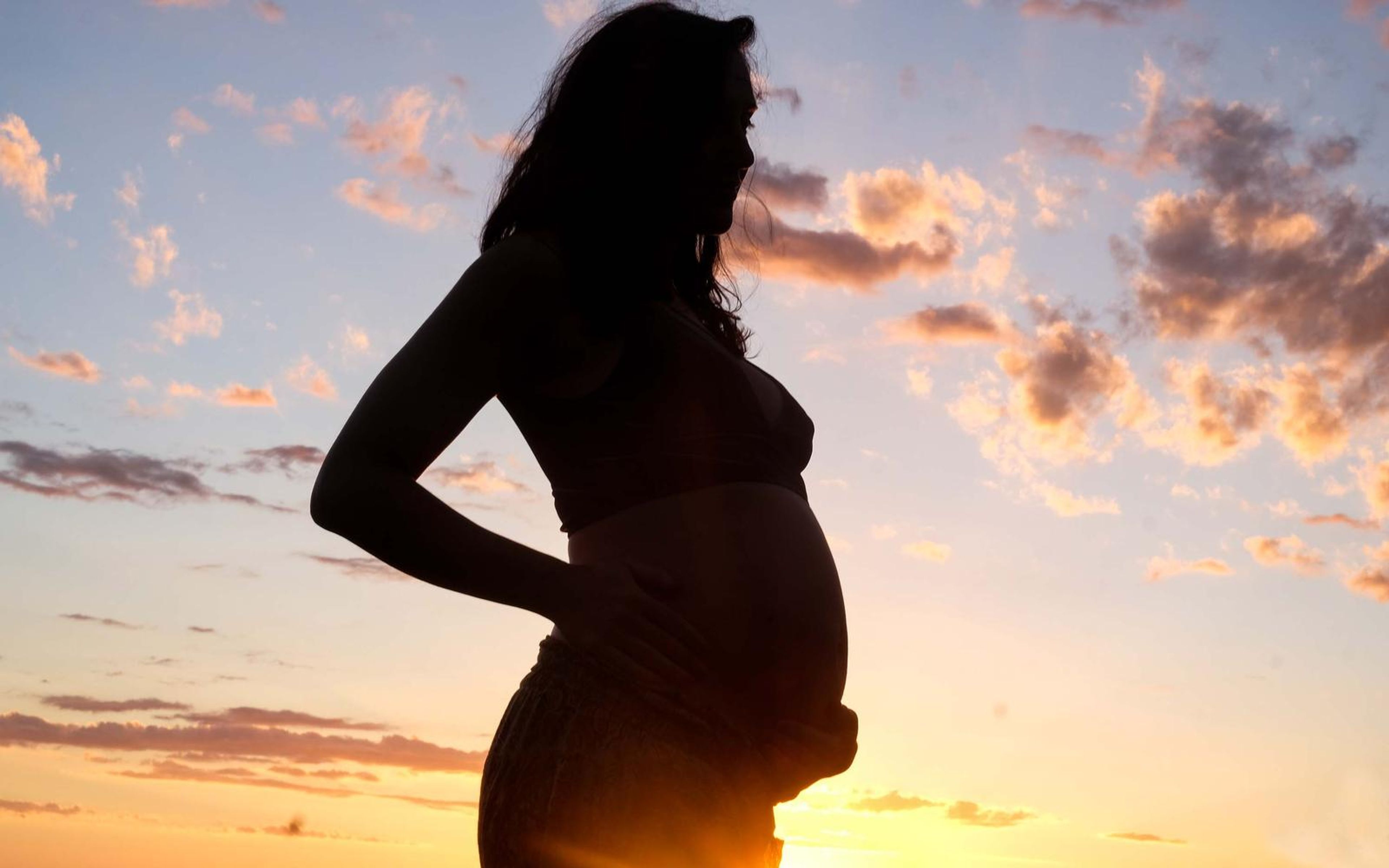 Silhouette of a pregnant woman in profile at sunset, hand on her lower back, scattered pink clouds