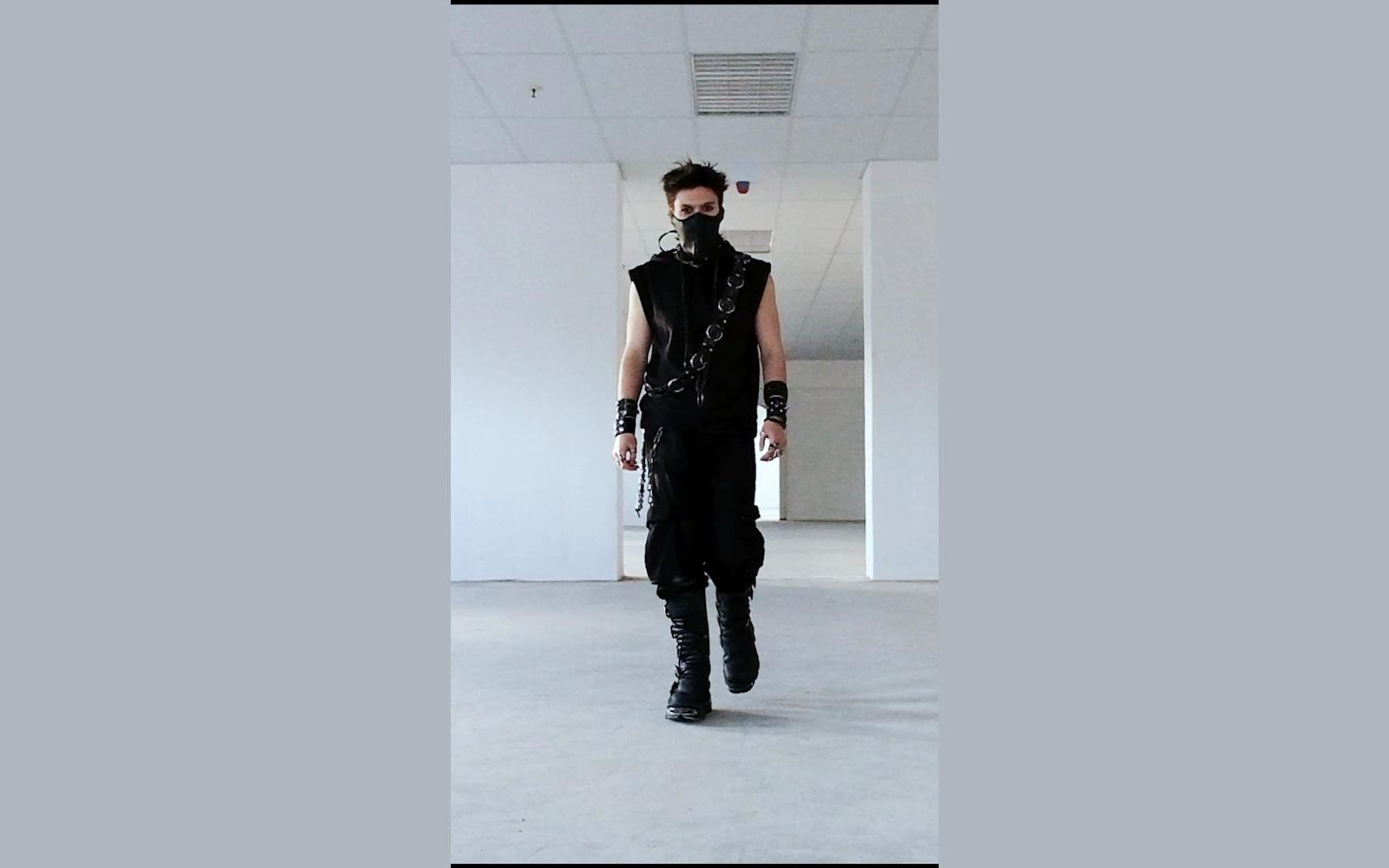 Masked figure walking forward in a sleeveless black outfit with wrist cuffs and a chain across the chest