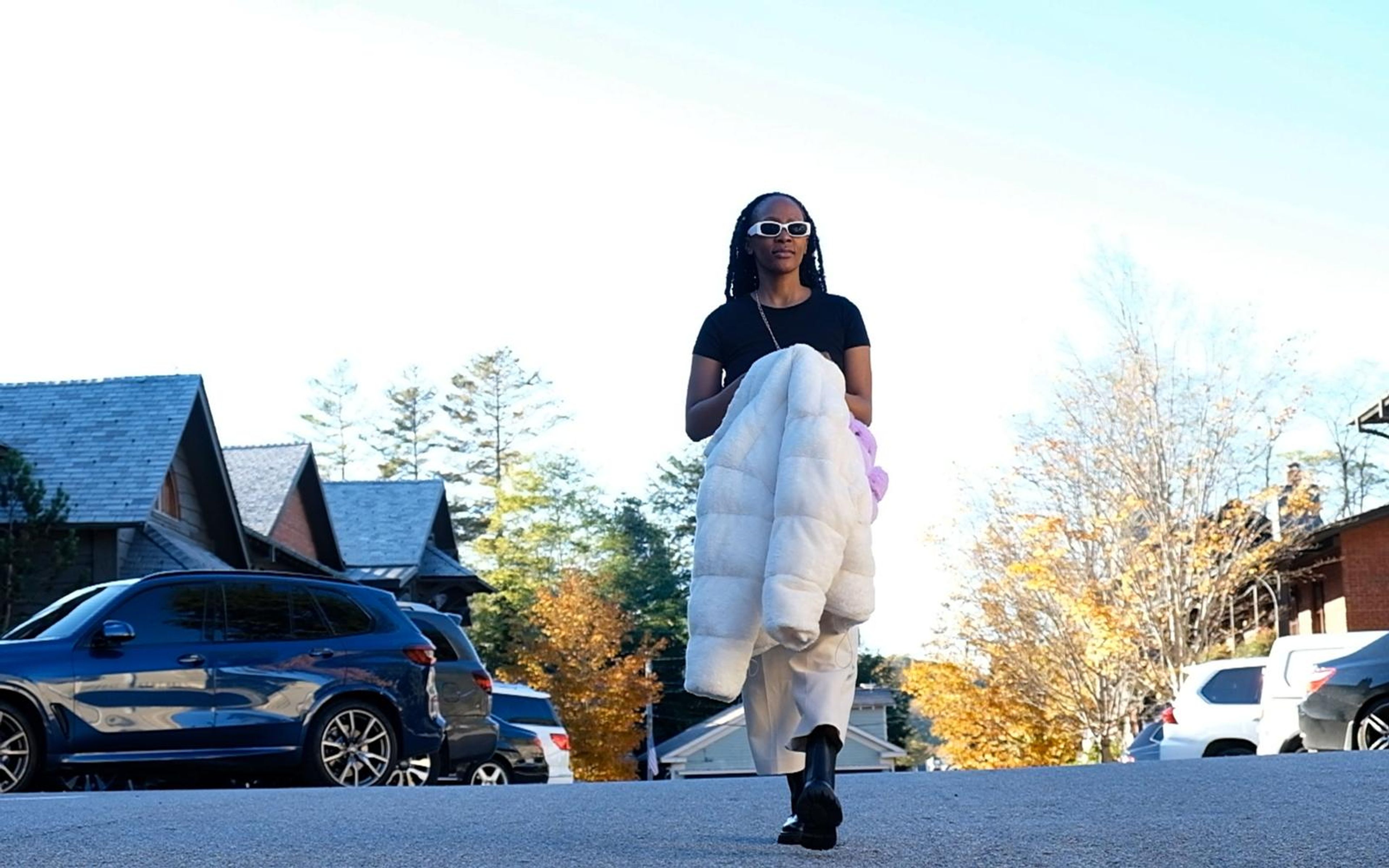 Model walking toward camera holding the white fur jacket across her arm, blue sky and rooftops
