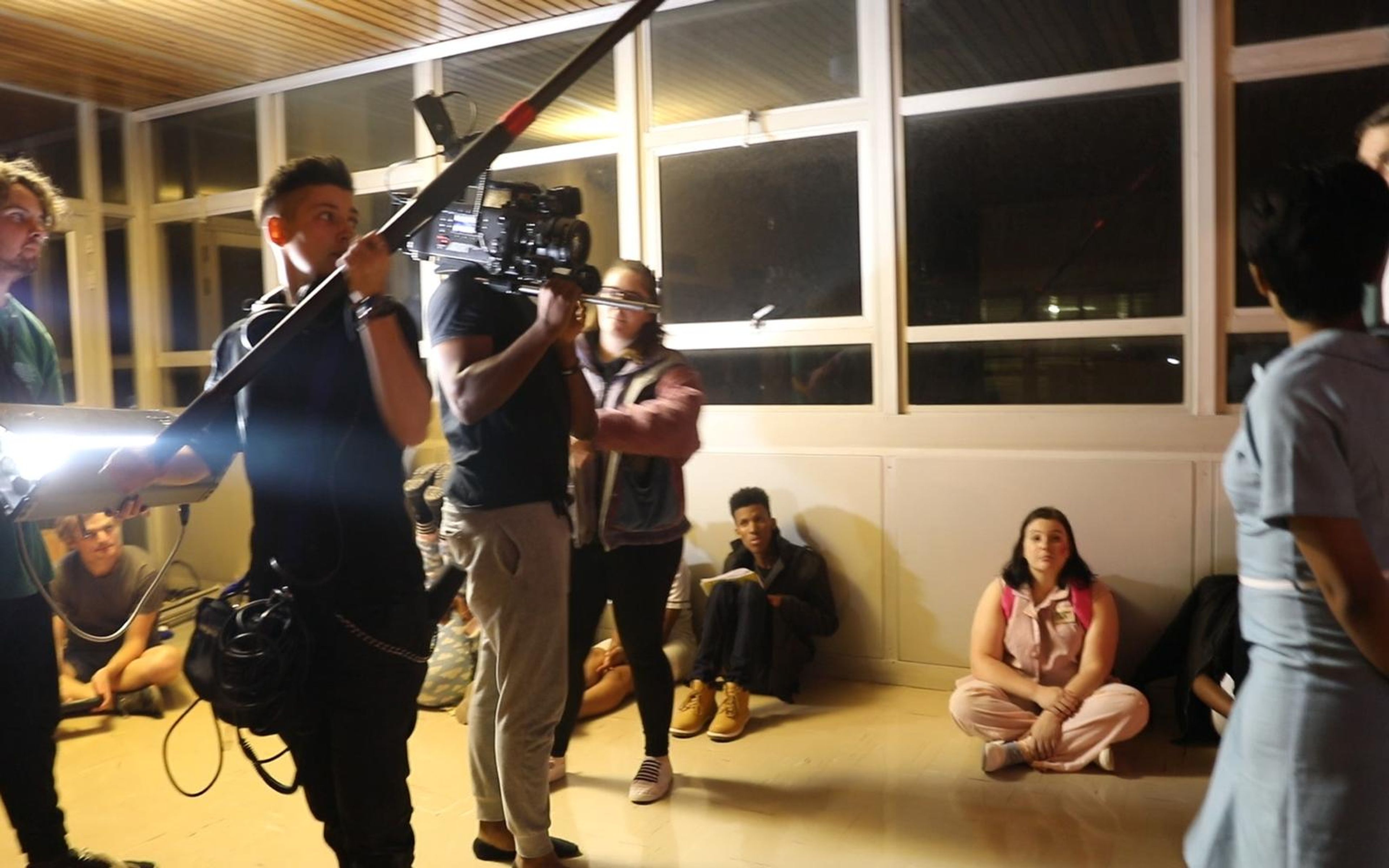 Boom operator and camera crew working in a warm-lit lounge while cast members sit on the floor