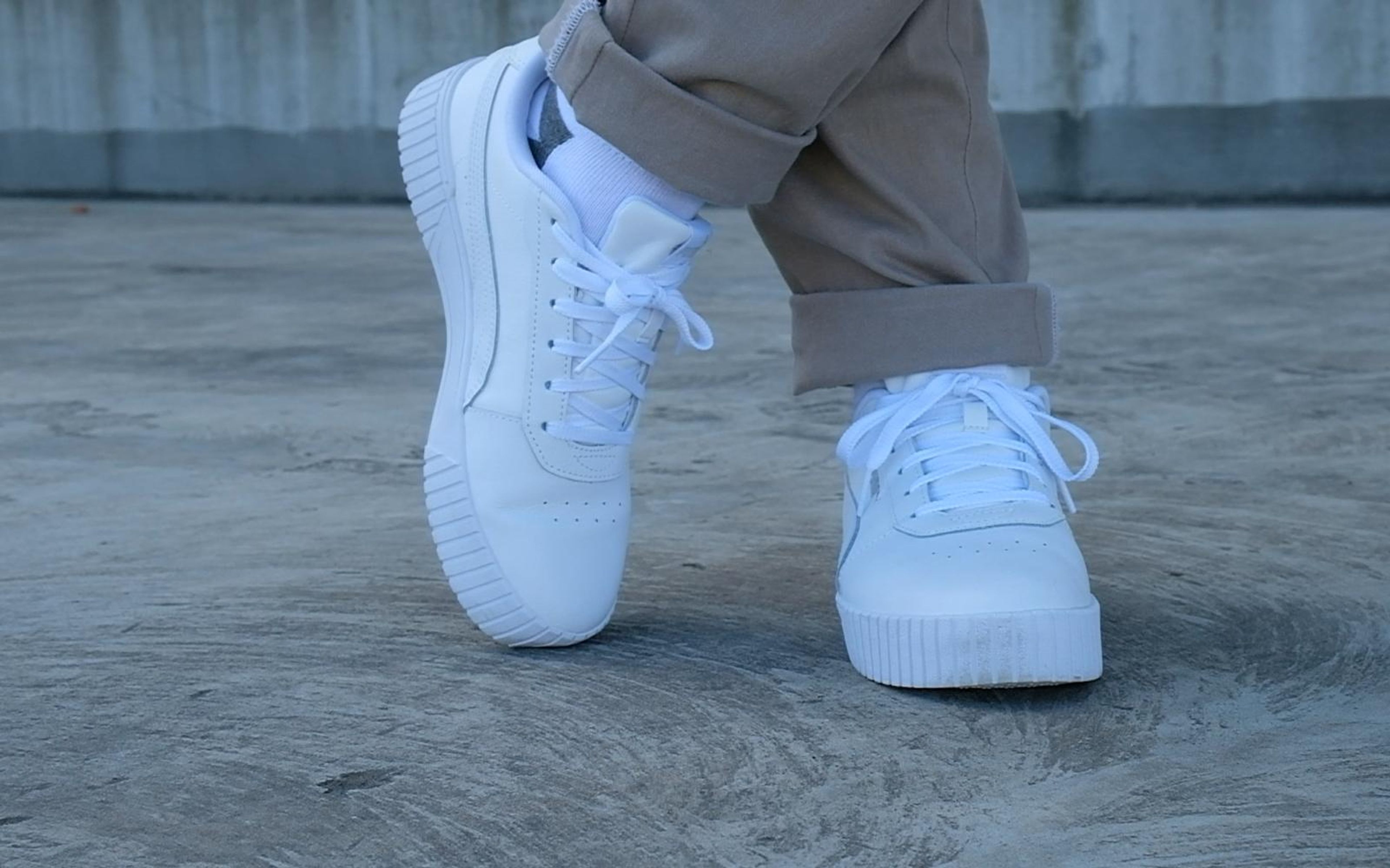 Side view of white sneakers, one tipped on its sole edge, tan trousers, muted blue concrete
