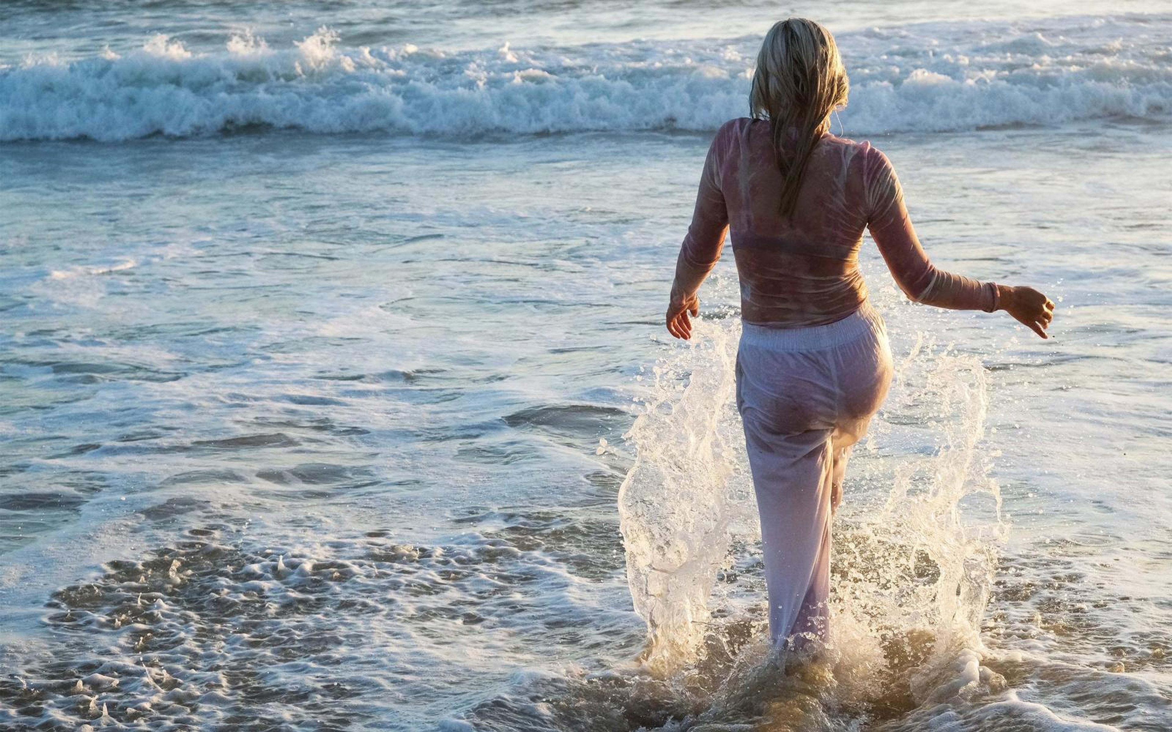 Woman seen from behind running through the surf in white trousers and a wet pink top, water splashing around her legs