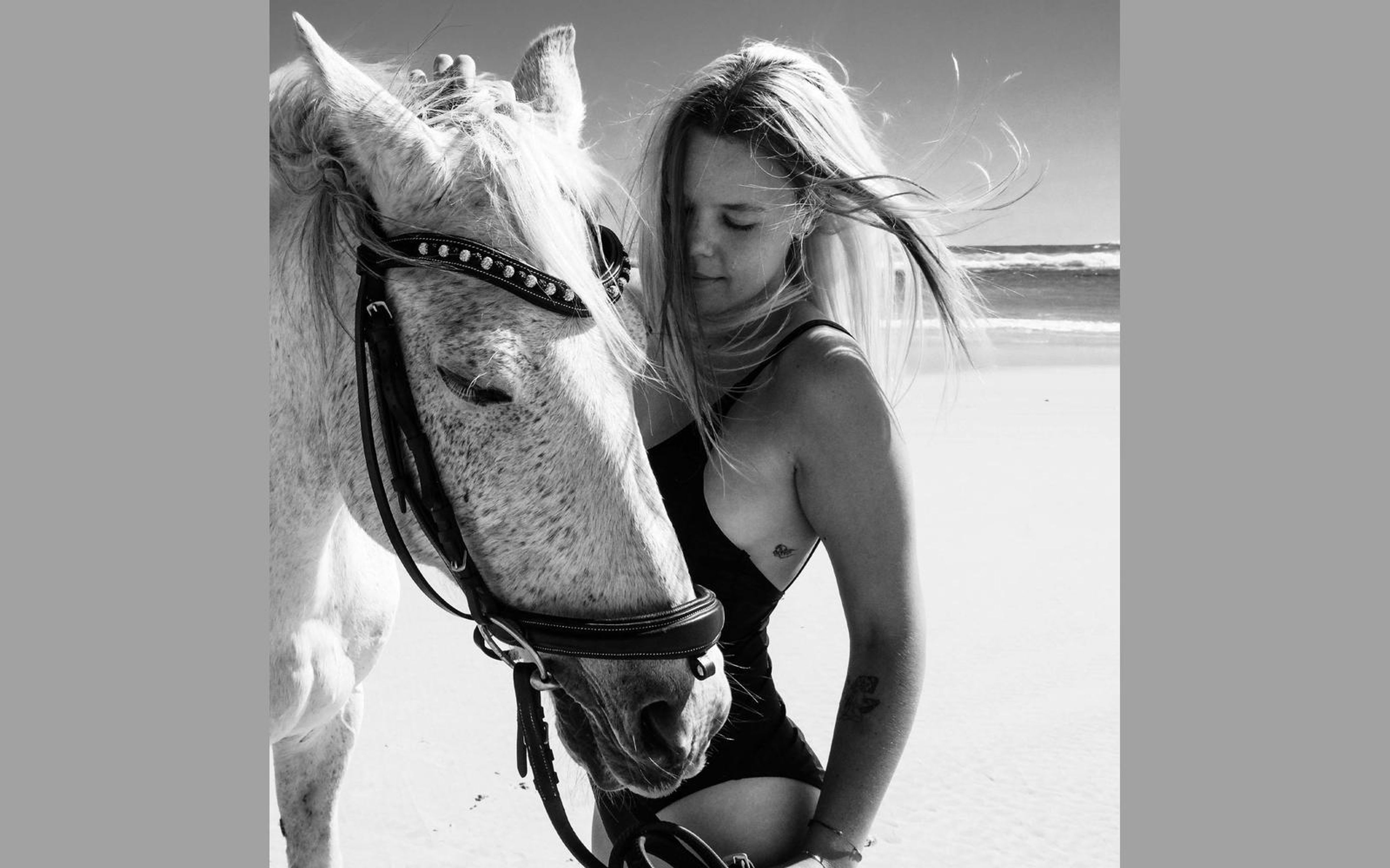 Black and white version of a woman in a swimsuit resting her head against a horse face with bridle, windblown hair, beach behind